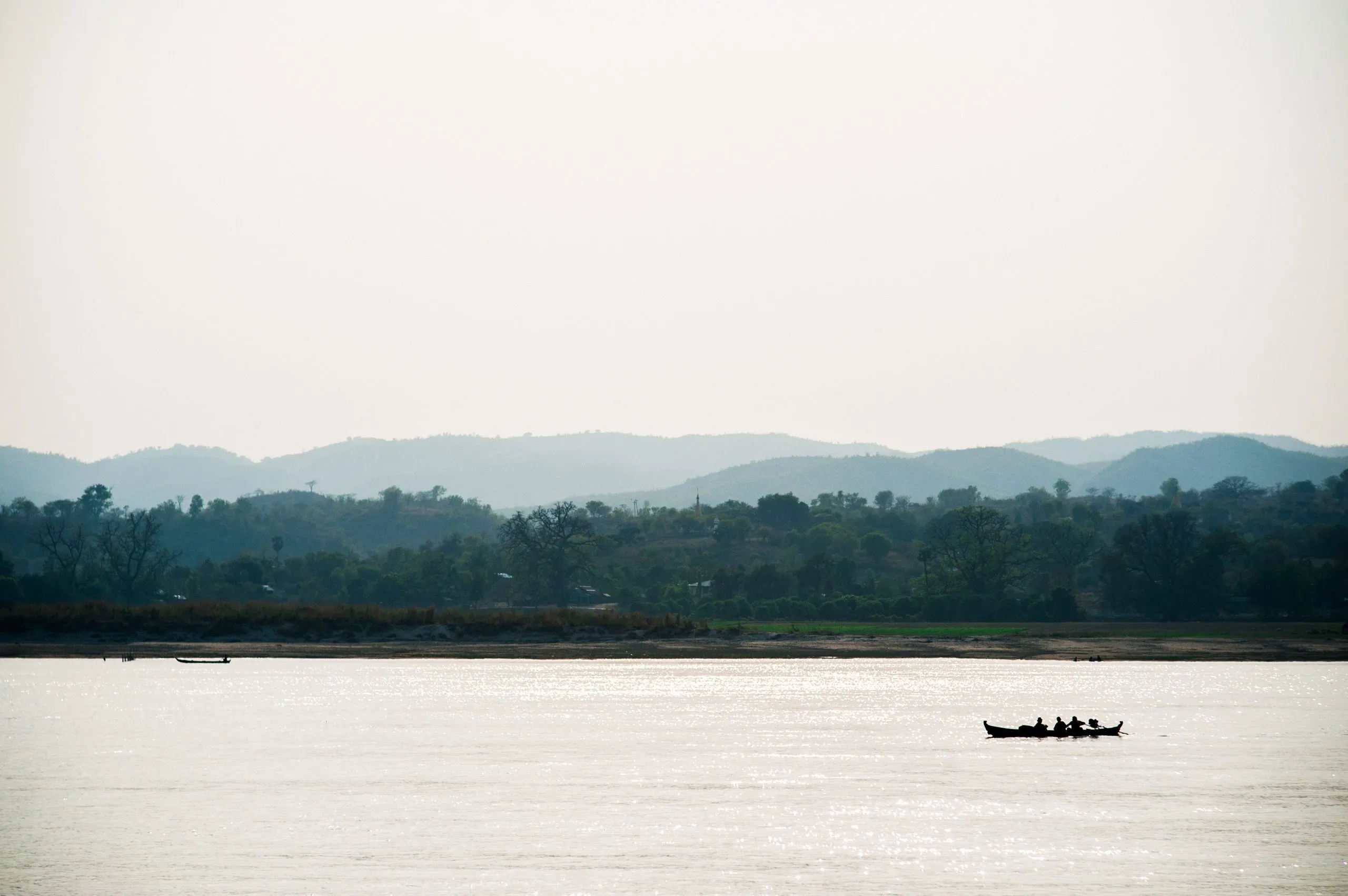 A small boat crosses the misty Irrawaddy River, with hazy hills rising behind, in central Burma.