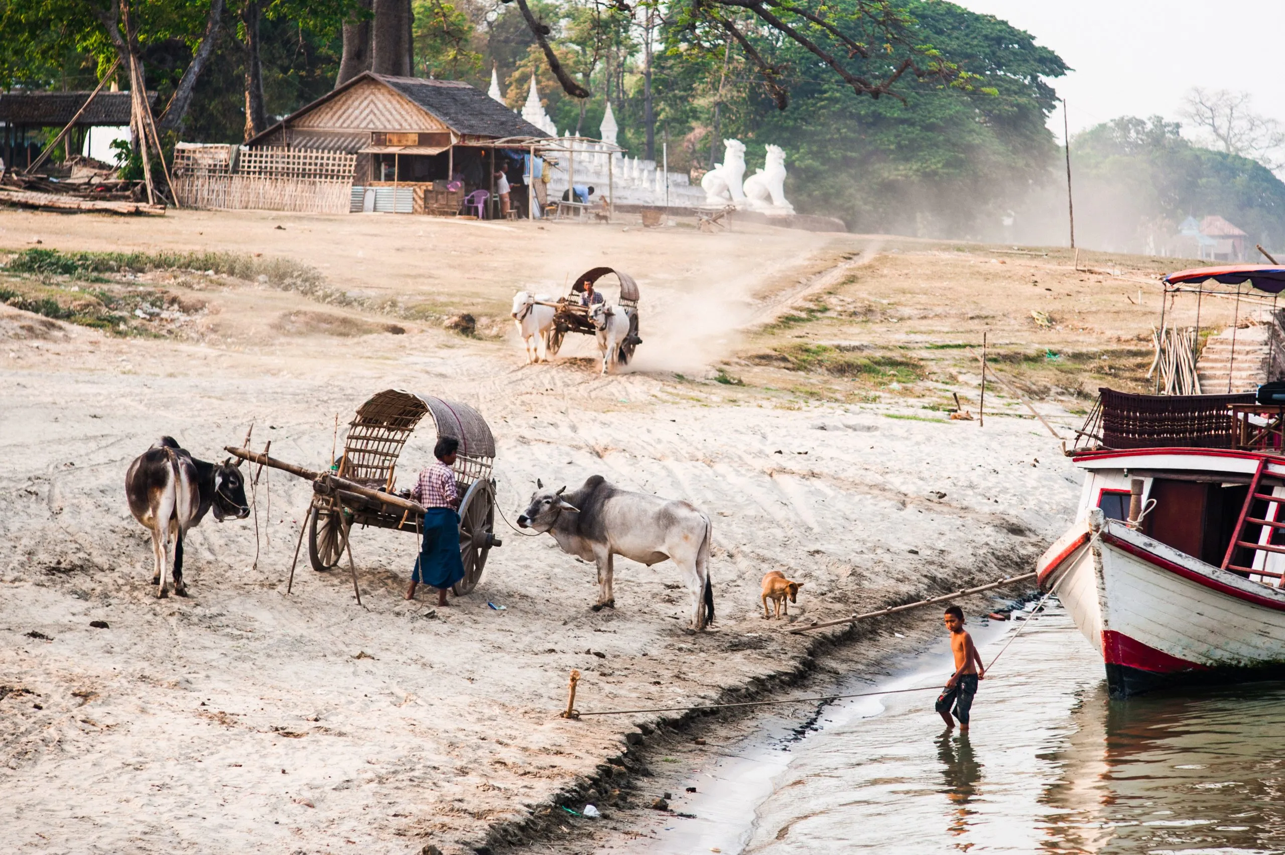Wooden oxcarts stand on a sandy riverbank below a Burmese pagoda, with a boat moored in the river.