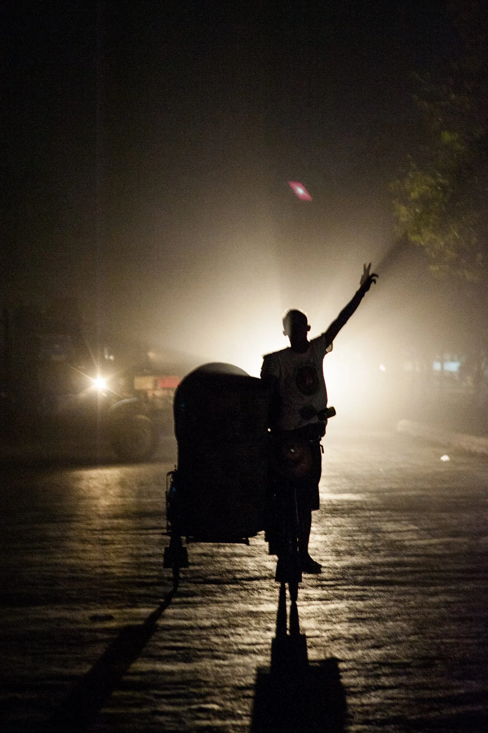 A rickshaw driver stands and raises his arm on a misty night street in Mandalay, Burma.