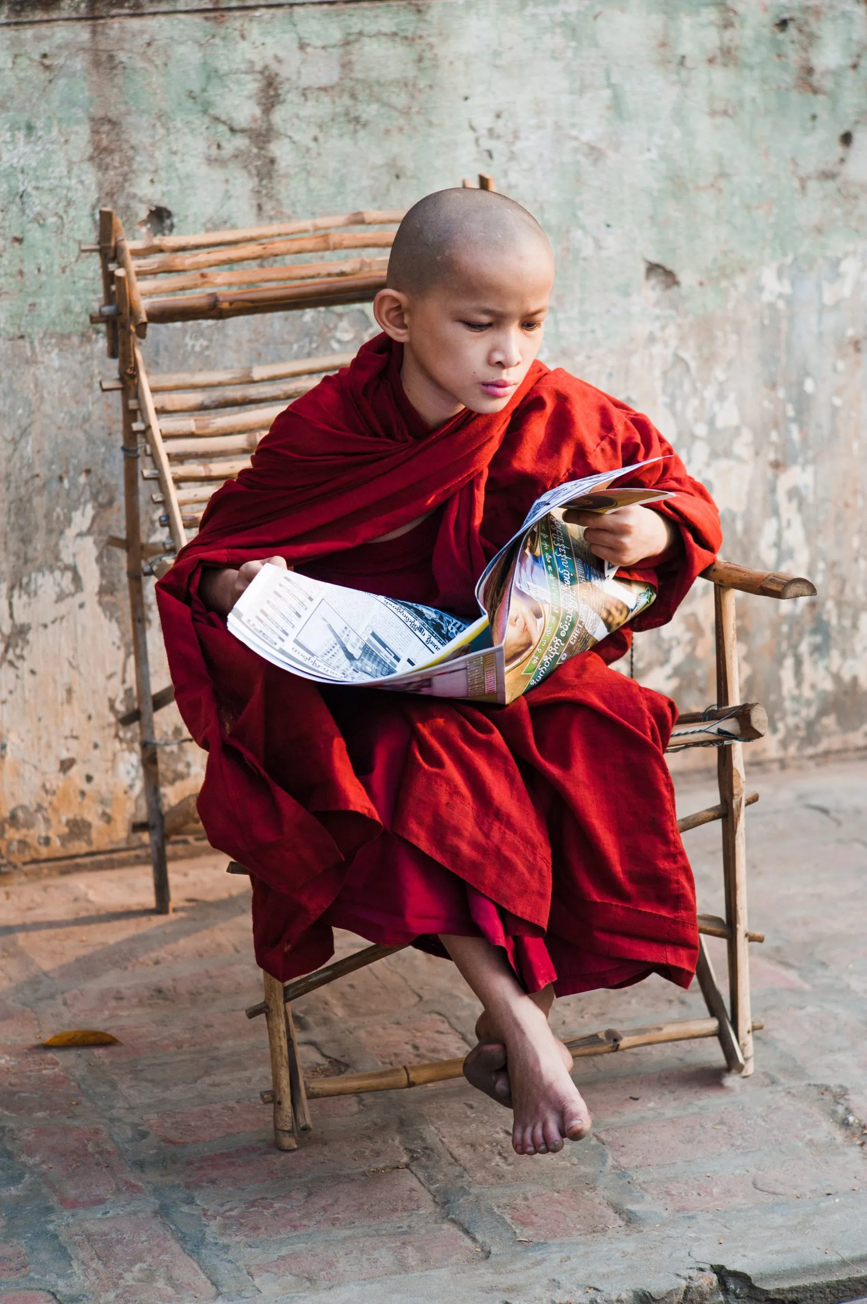 A young monk reads a magazine while seated cross-legged on a bamboo chair in Burma.