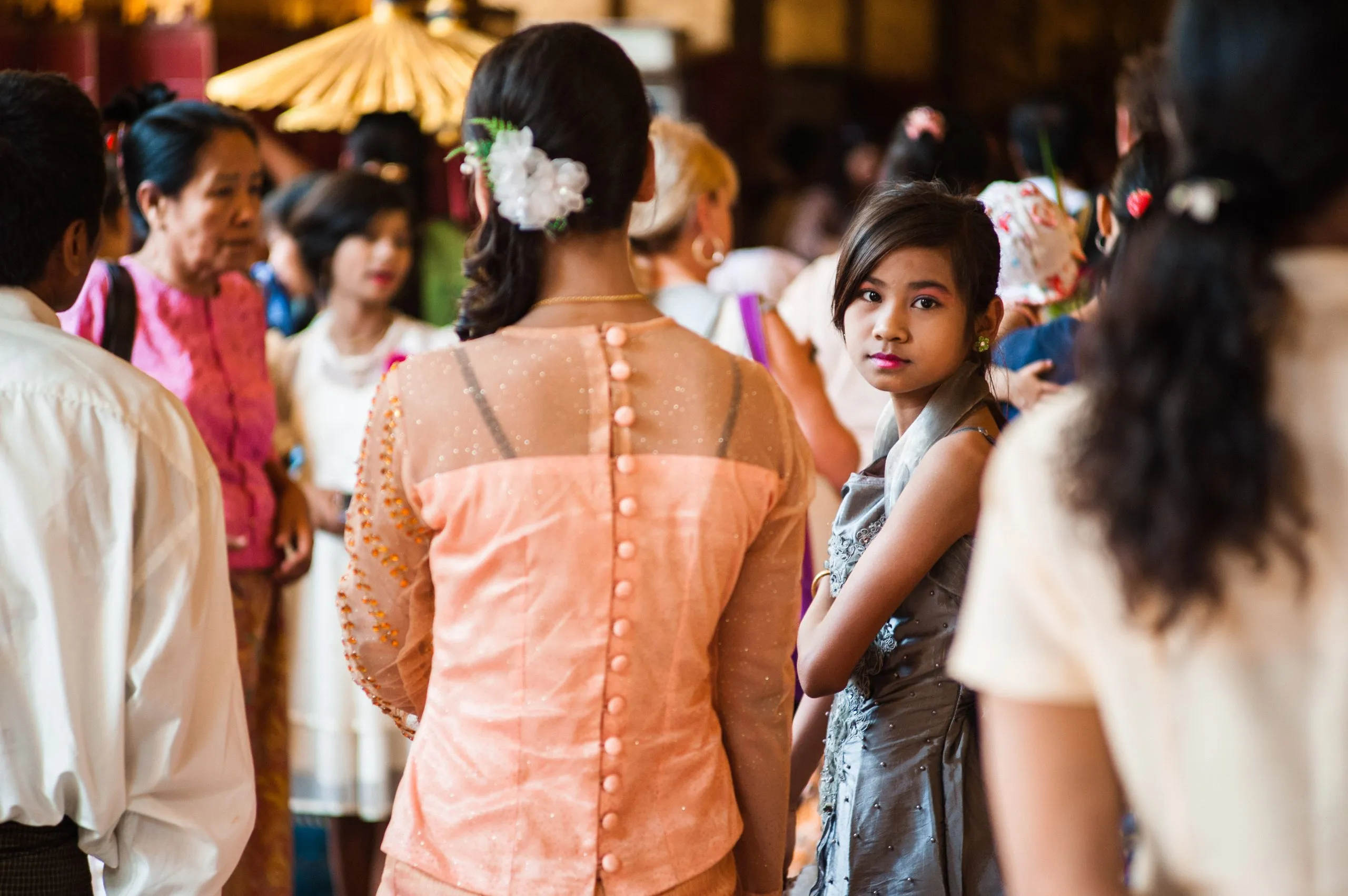 Women in traditional Burmese formal dress gather in a hall, with a parasol in the background.