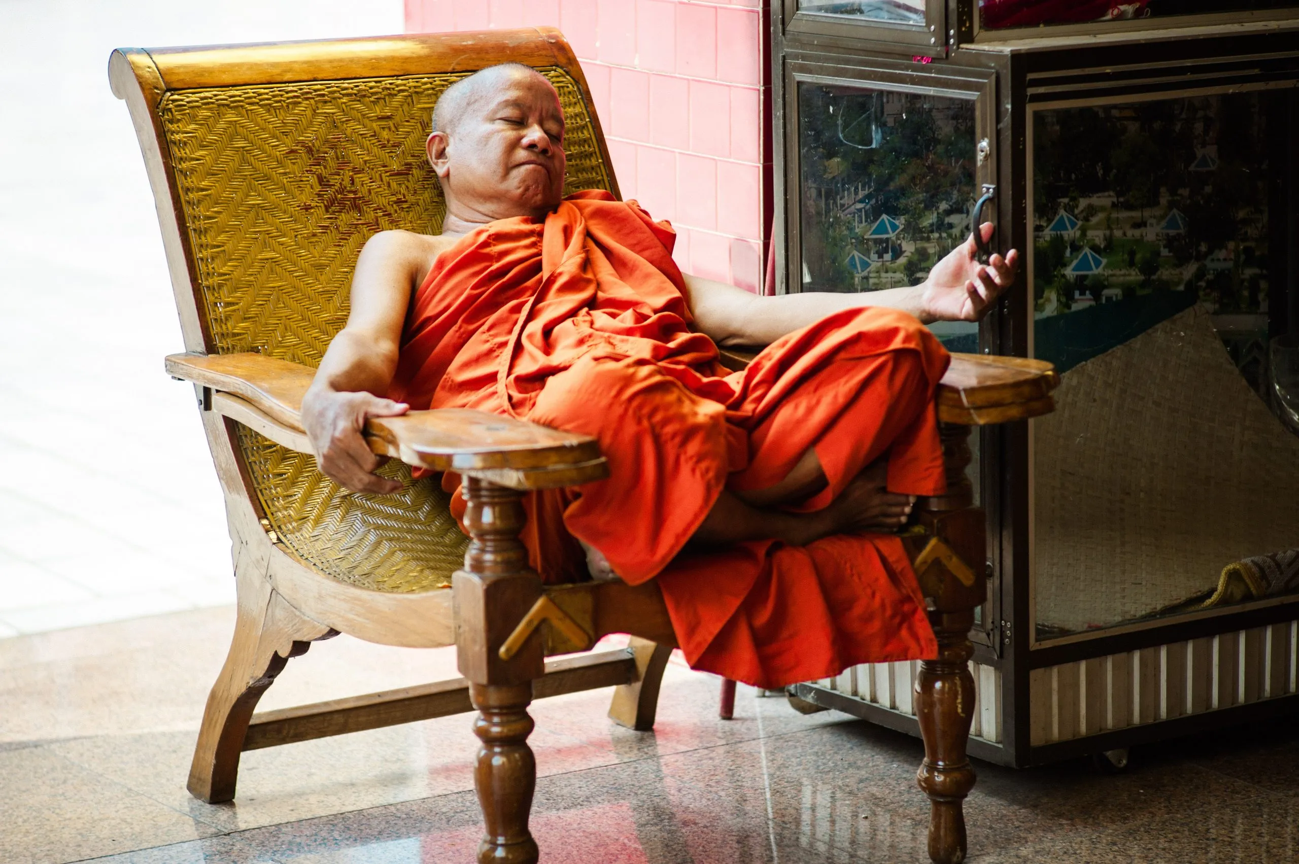 An older monk in saffron robes reclines in a wooden chair inside a Burmese monastery.