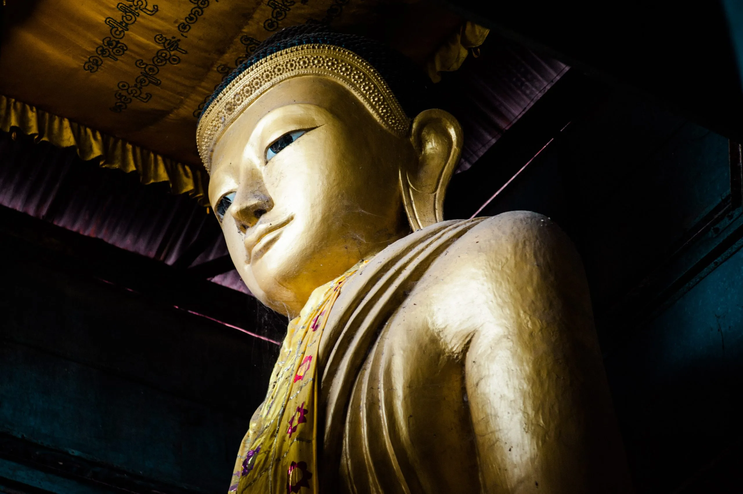 A close detail of a large gilded Buddha statue under the painted ceiling of a Burmese temple.