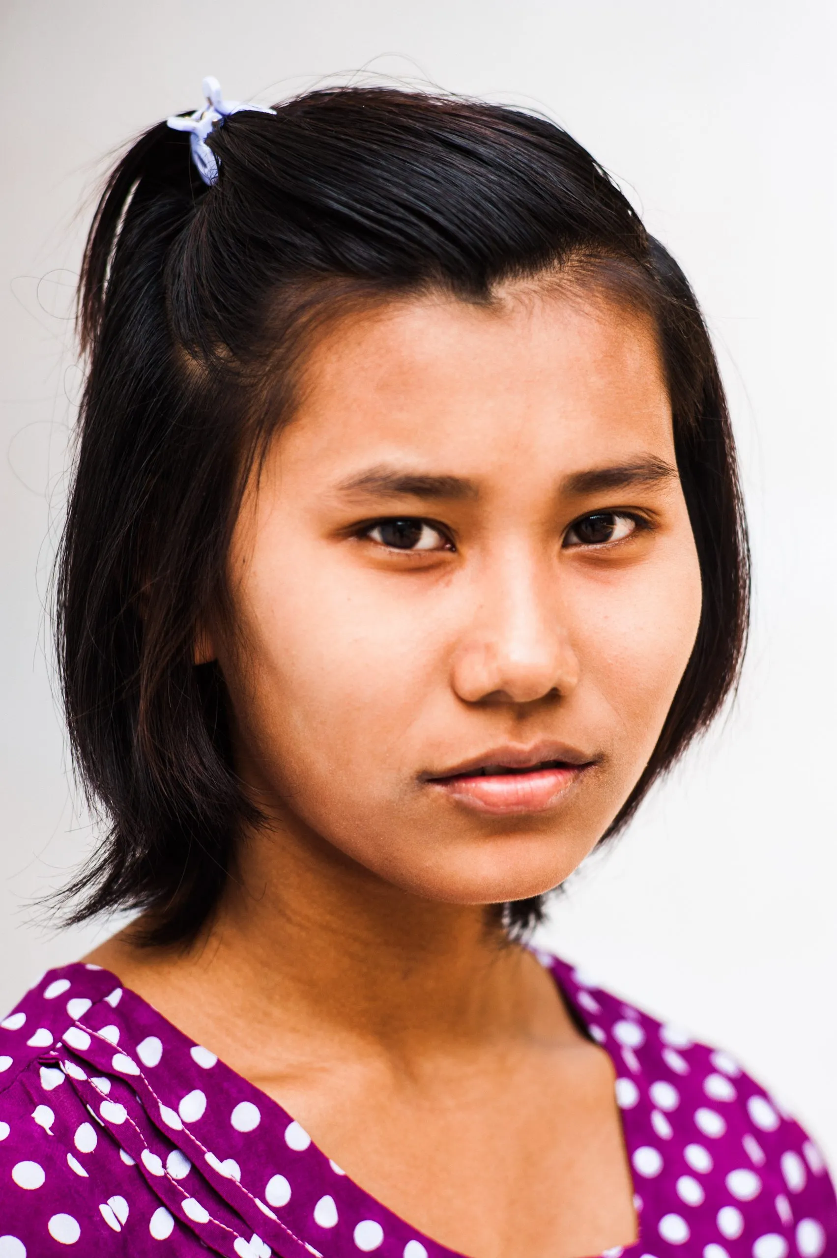 A close portrait of a young Burmese woman in a purple polka-dot top against a pale wall.