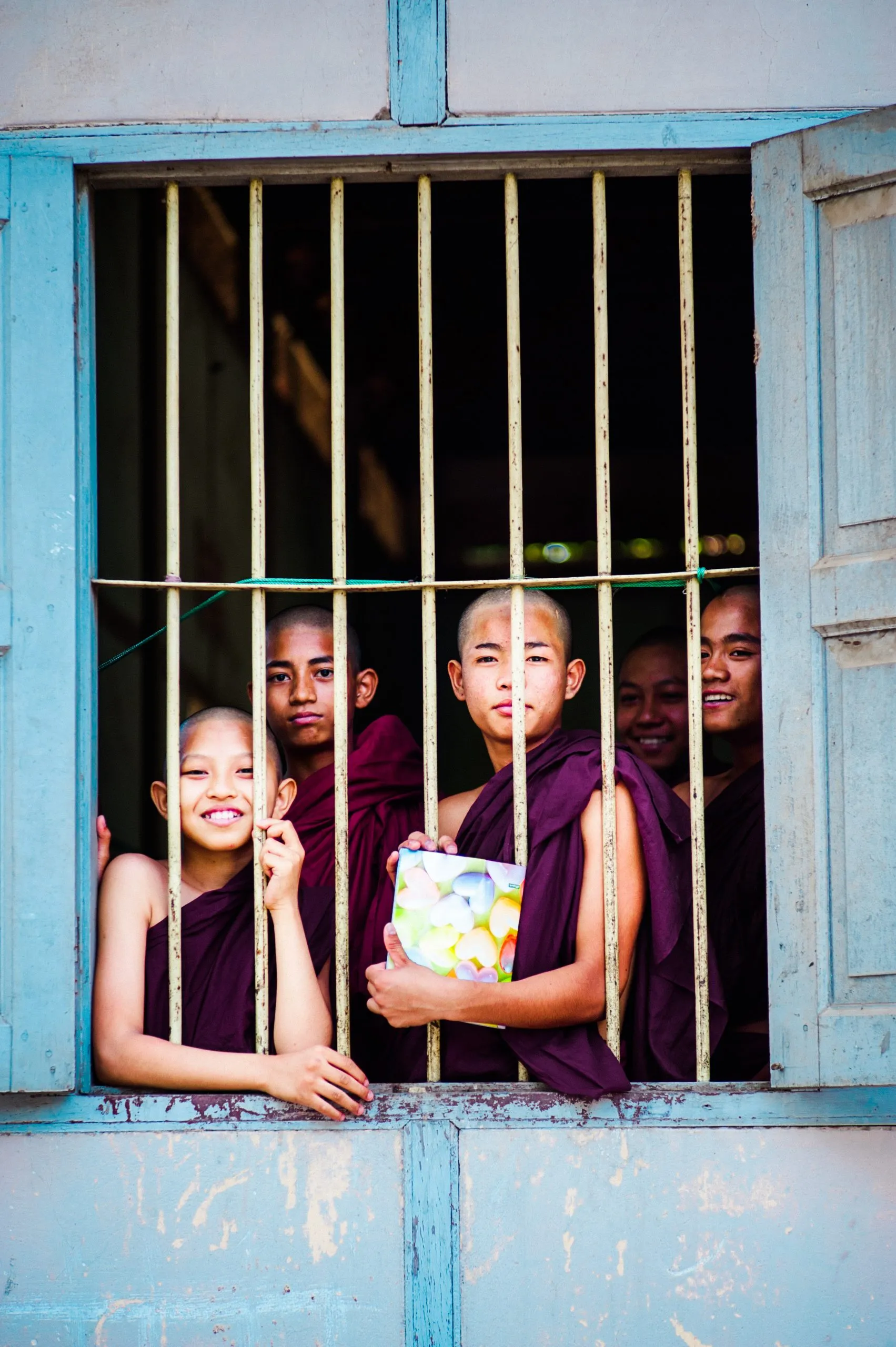 A group of young monks smile through the blue-painted bars of a monastery window in Burma.