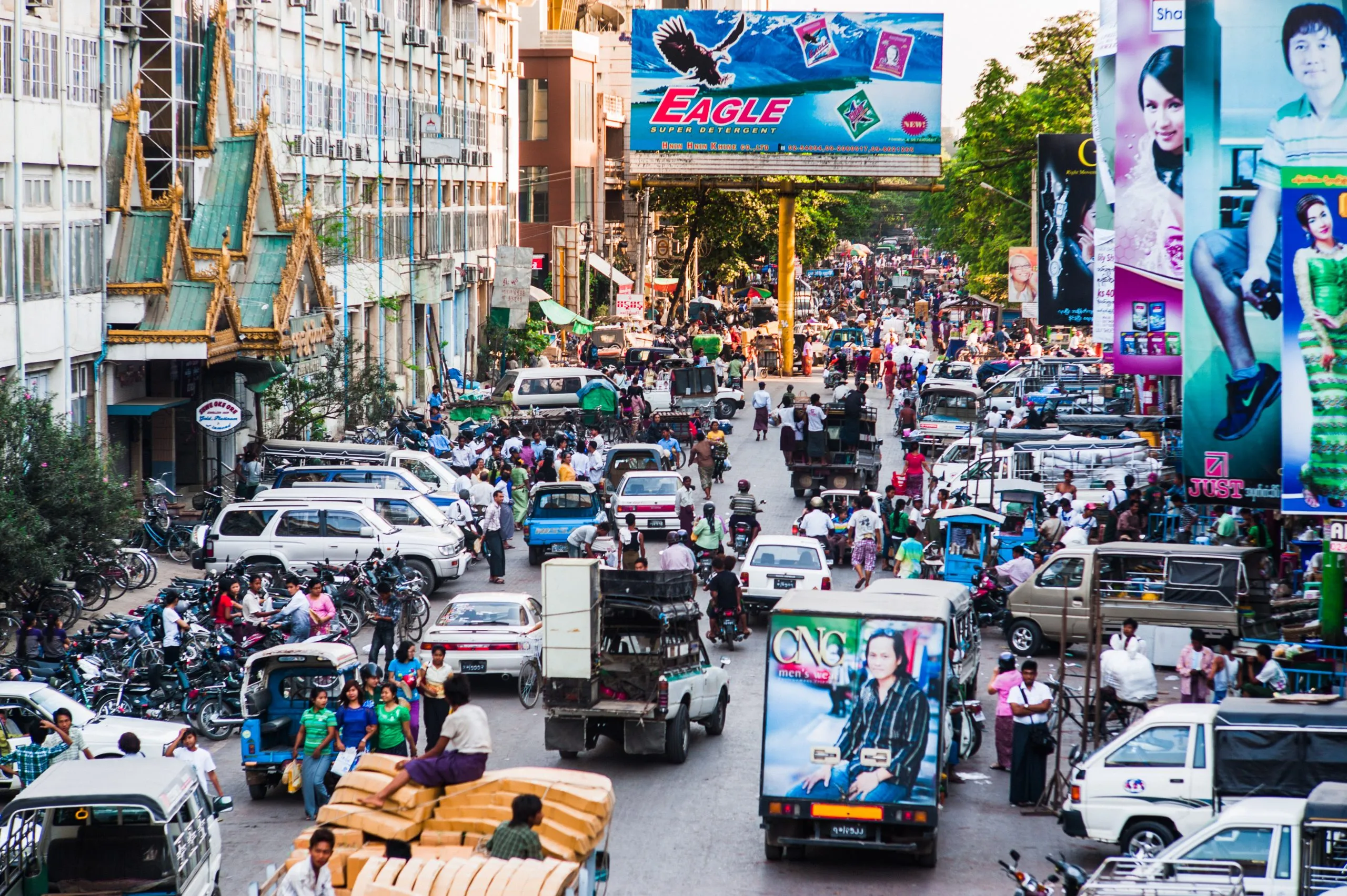 A bustling Mandalay street choked with traffic, beneath a large Eagle advertising billboard.