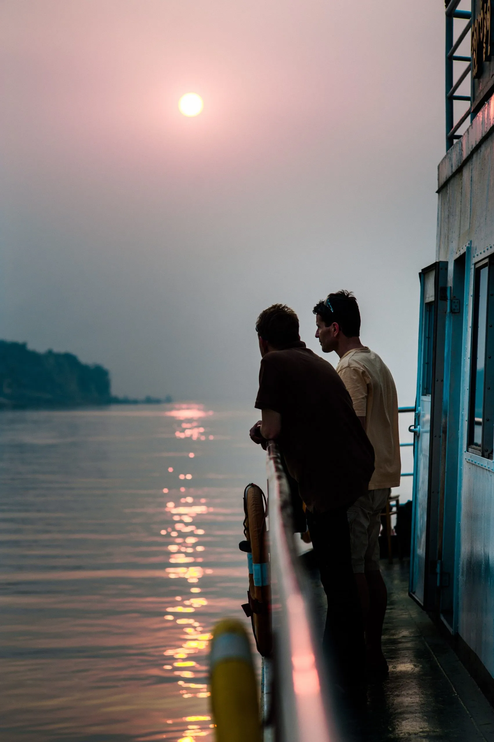 Two travellers on the deck of a boat watch a soft pink sunset reflected on the Irrawaddy River.