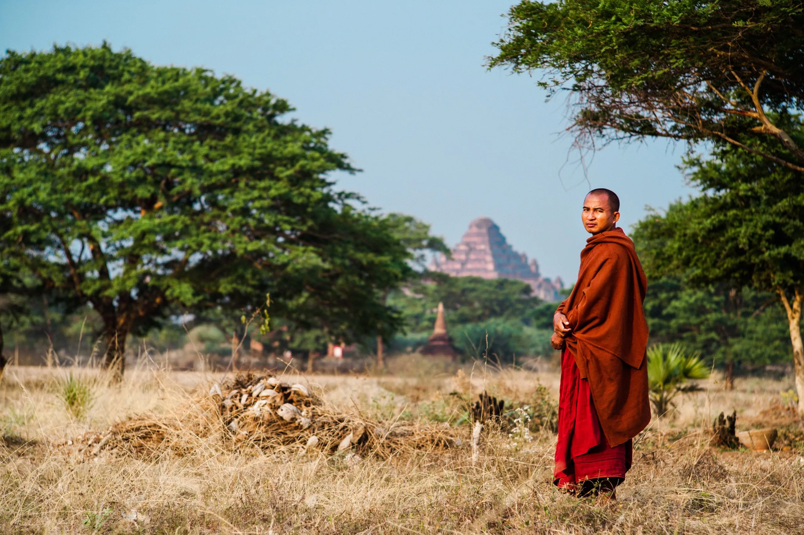 A monk in red robes pauses in a dry Bagan field, with a brick stupa visible behind him.