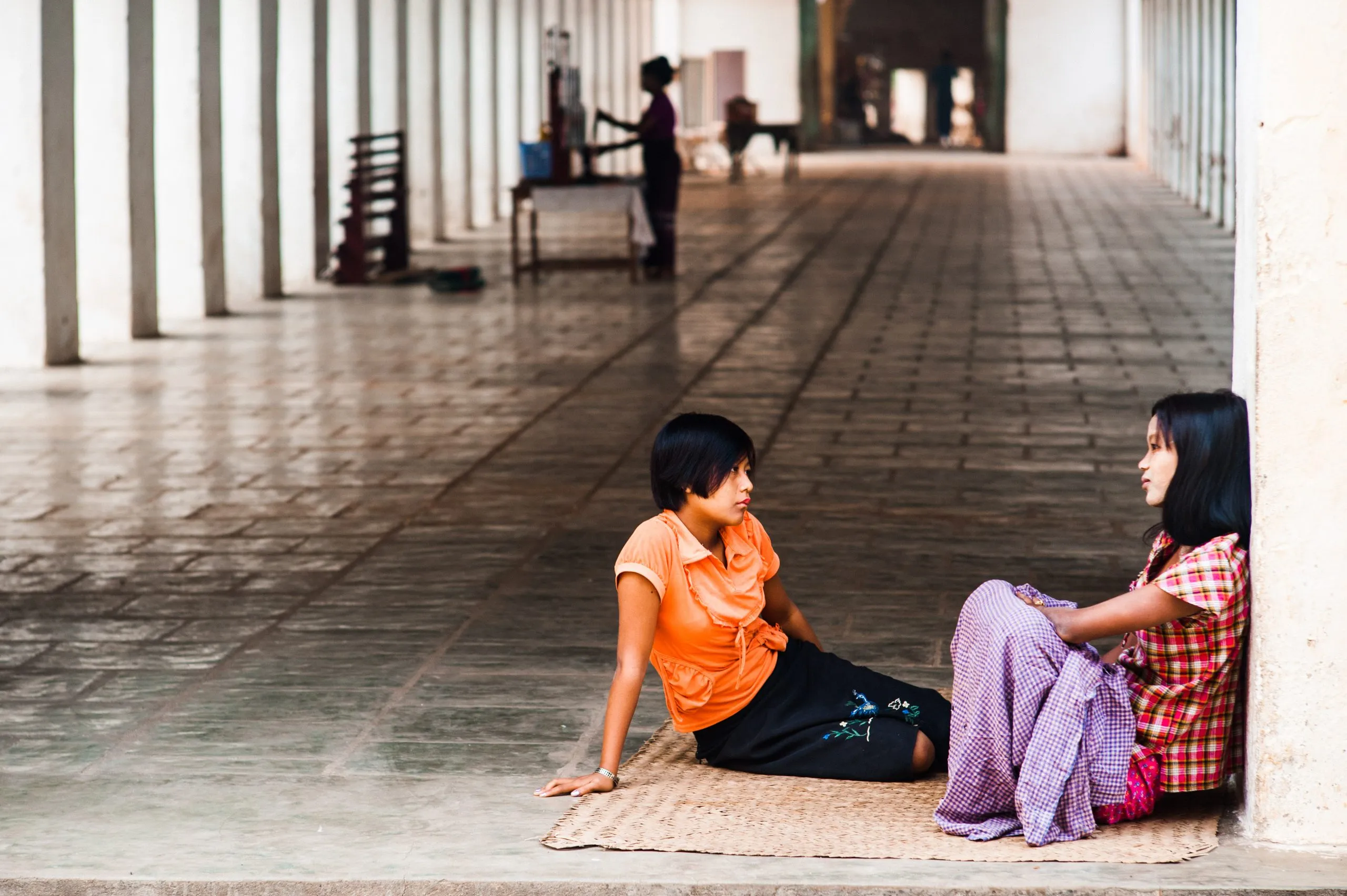 Two young women sit talking on the floor of a long covered walkway in a Burmese monastery.