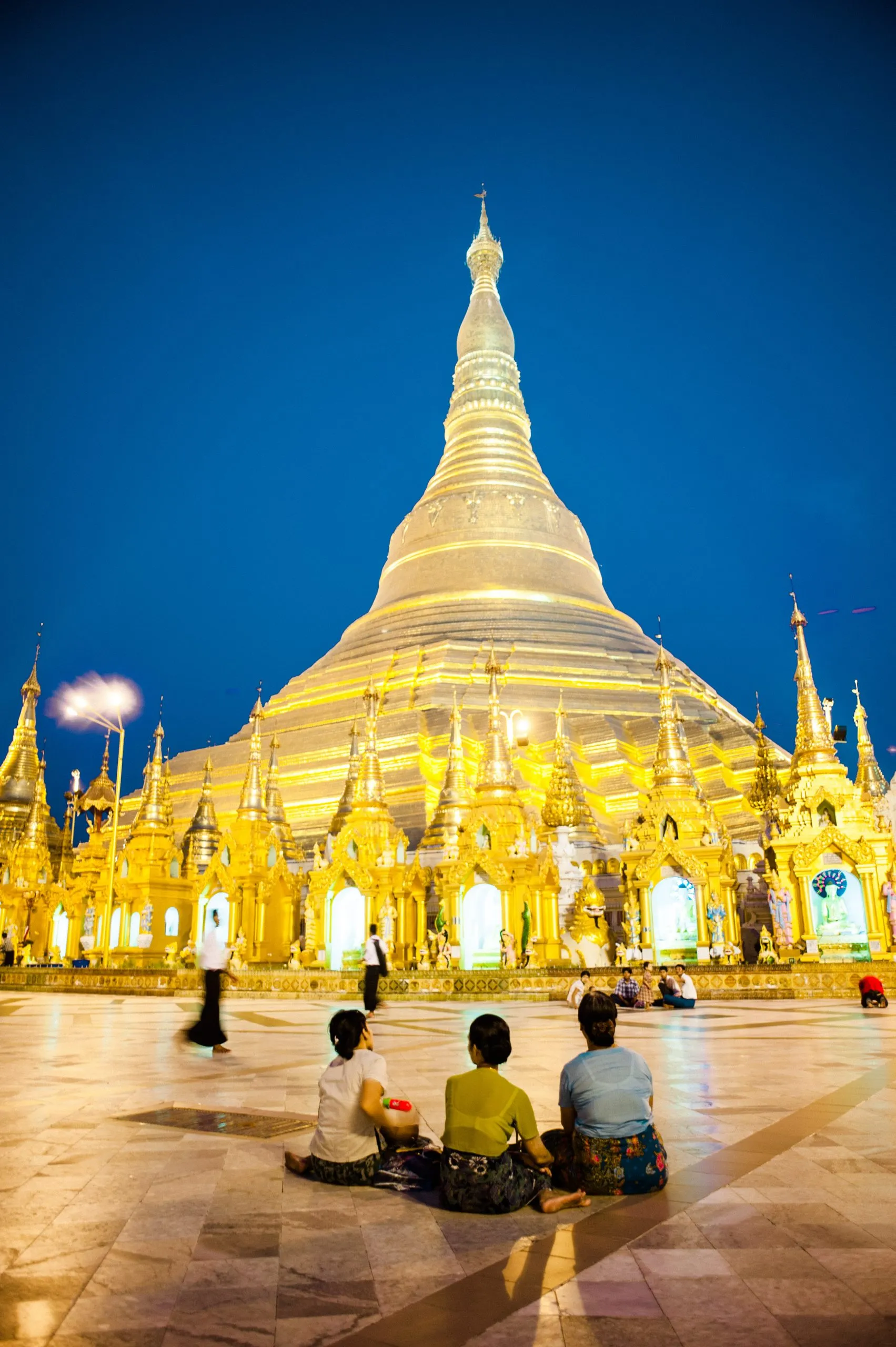 Three people sit before the Shwedagon Pagoda glowing gold against the deep blue night sky in Yangon.
