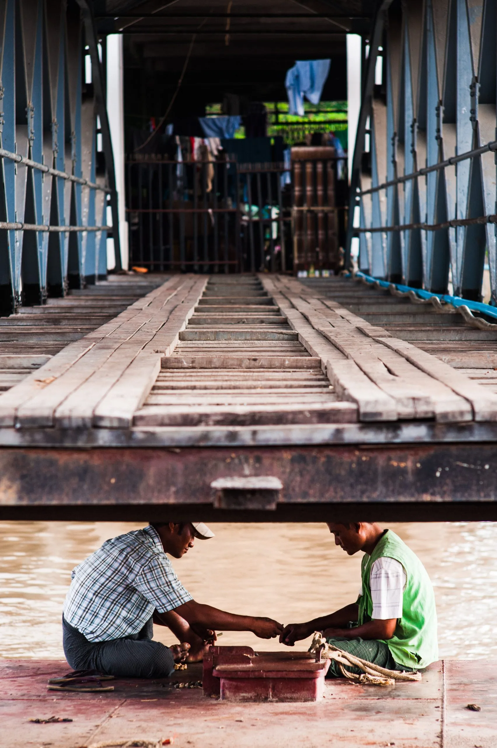 Two men sit beneath a wooden jetty, working with rope at the water's edge in Burma.