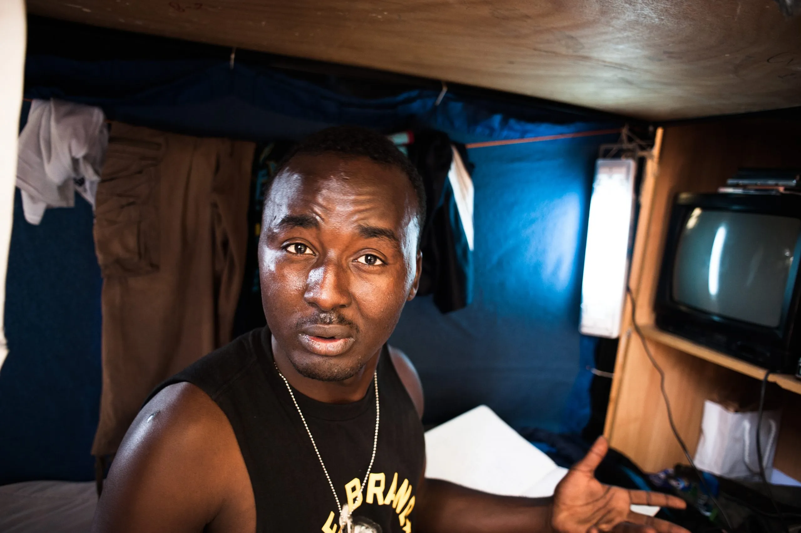 A man inside the curtained partition of his bunk, with a small television beside him, in the Malta housing hangar.