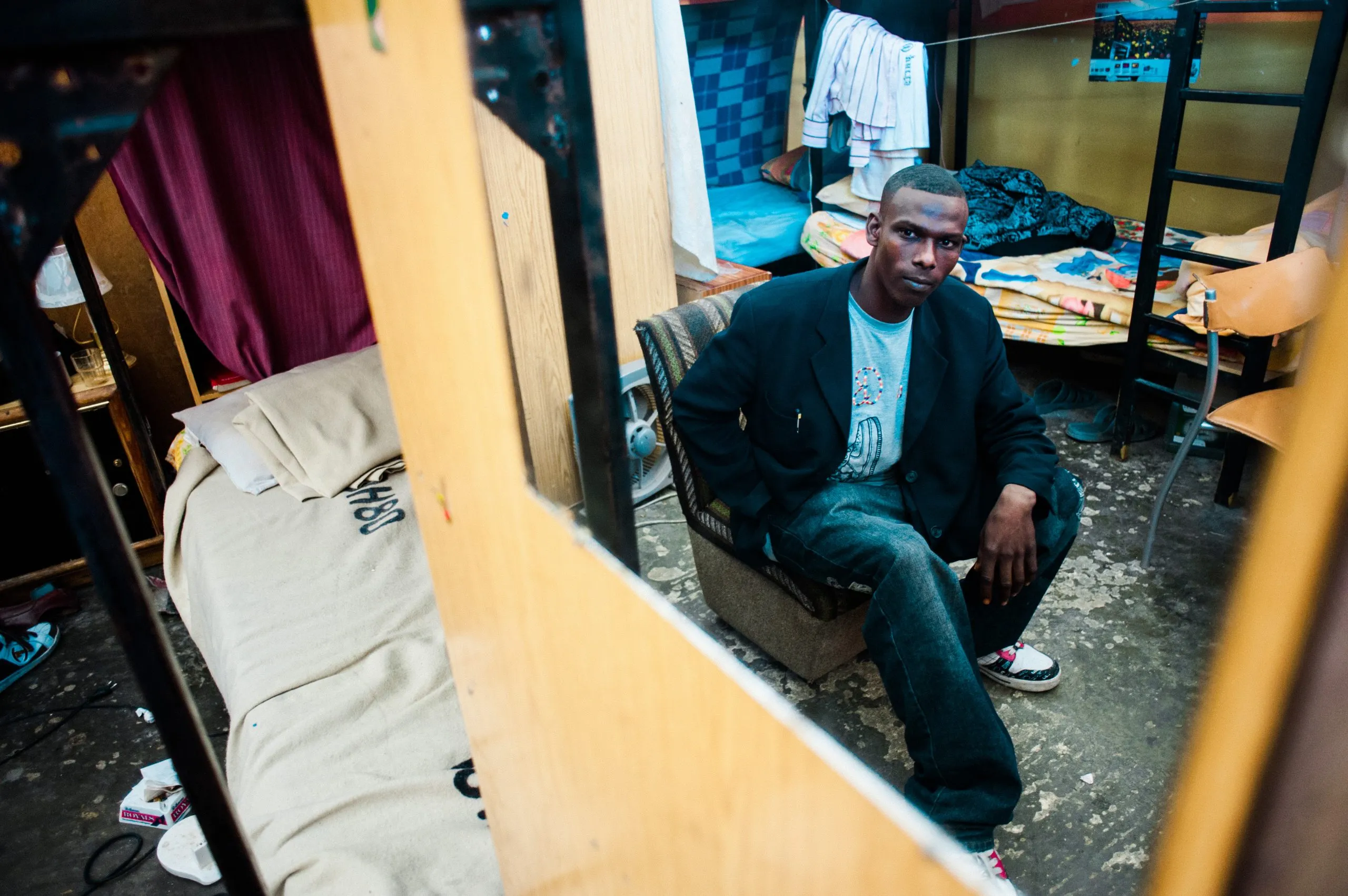 A man seated in a plastic chair, framed through the metal frame of a bunk bed in the Malta dormitory.