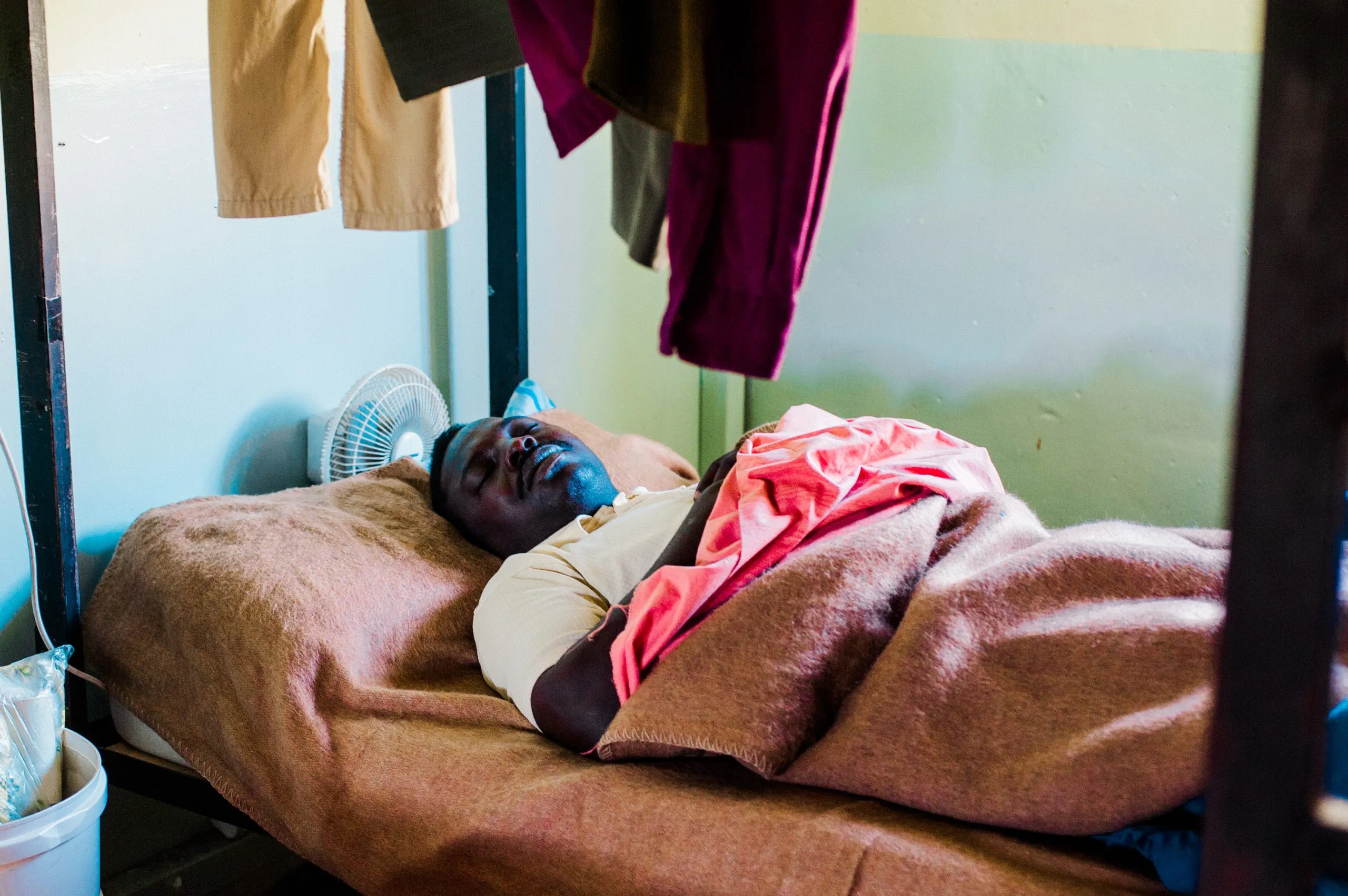 A man sleeps on a cot beside a small fan in the shared dormitory at the Malta housing hangar.