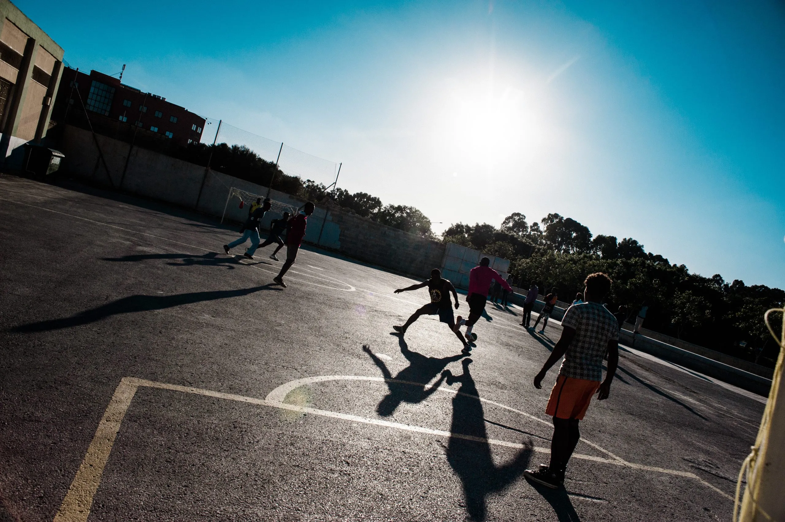 Silhouettes of people playing football on the outdoor court at the Malta housing compound, against a low sun.