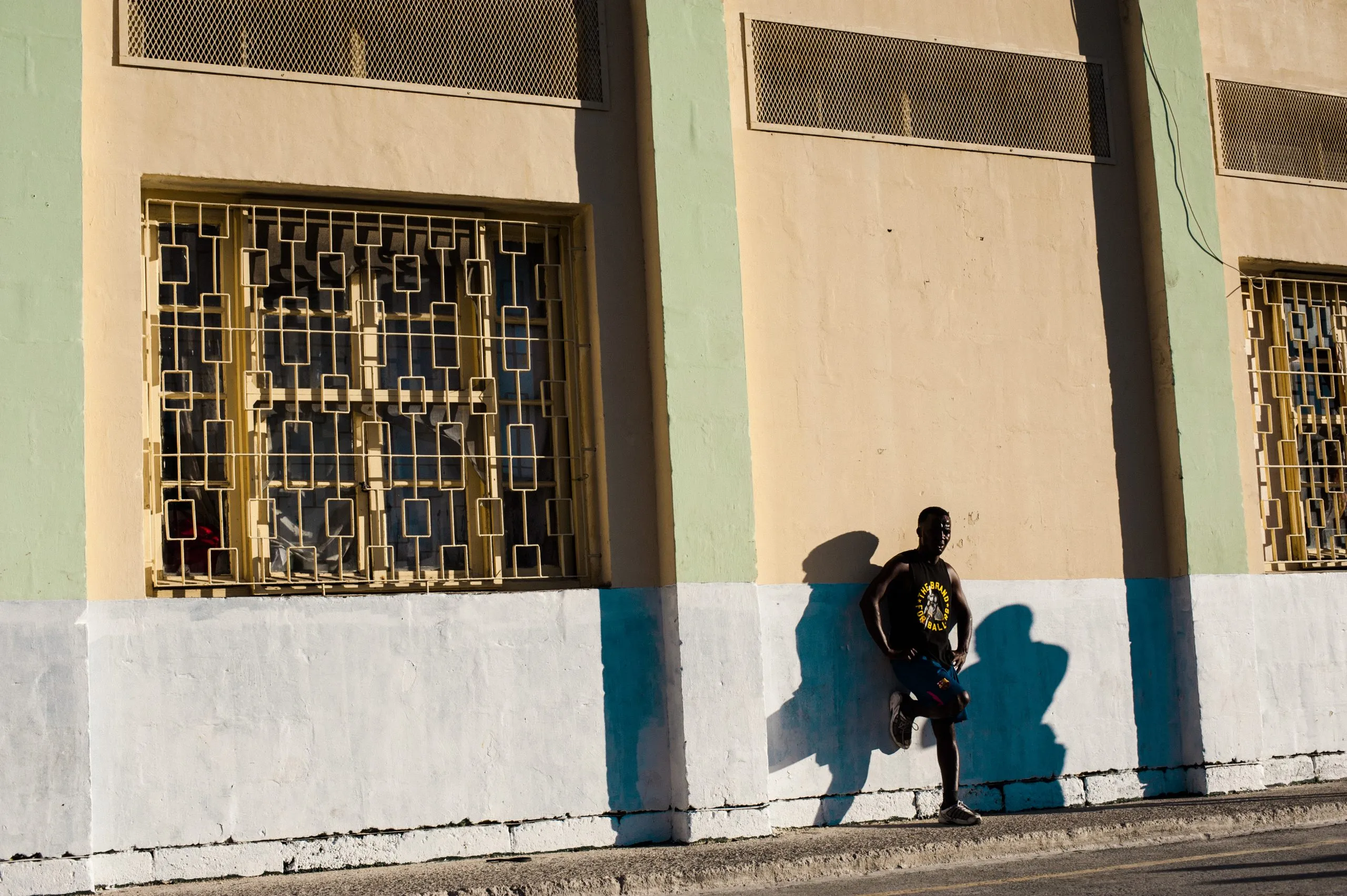 A man leans against the cage-windowed facade of a yellow building on a quiet street in Malta.