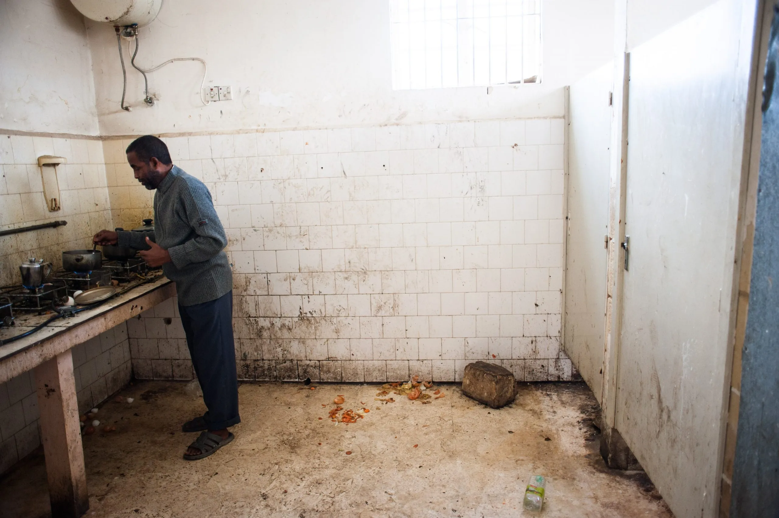 A man cooks at a wall of gas burners in a soiled tiled kitchen inside the Malta detention compound.