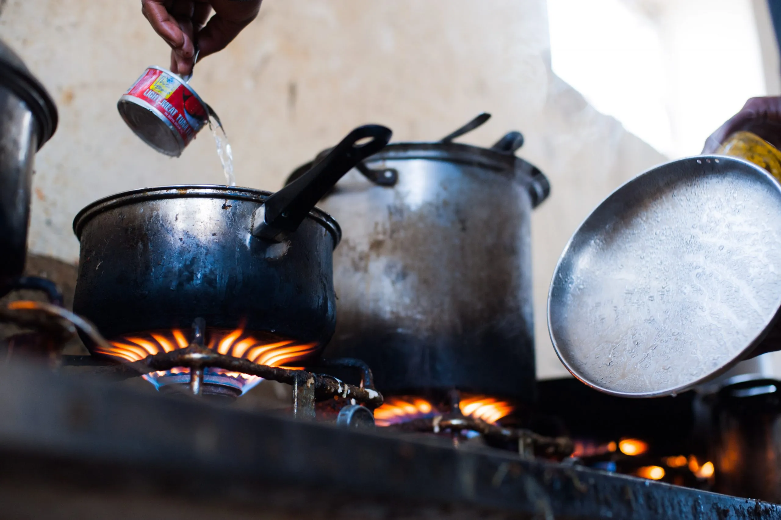 Hands cook over a gas burner with battered pots, inside the shared kitchen at the Malta compound.