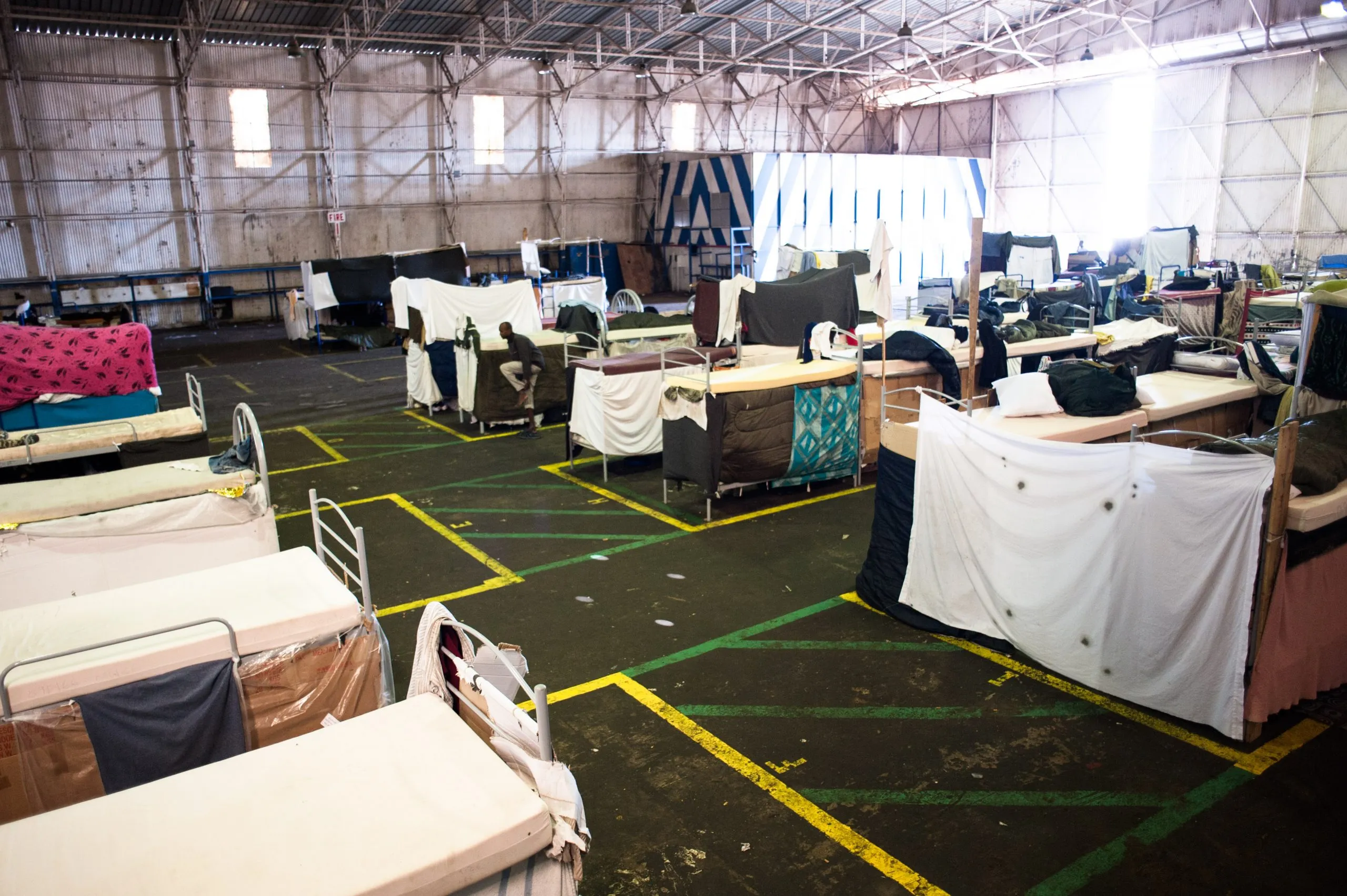 A wide view of a converted hangar in Malta where bunks are partitioned by hanging sheets above a yellow-gridded floor.