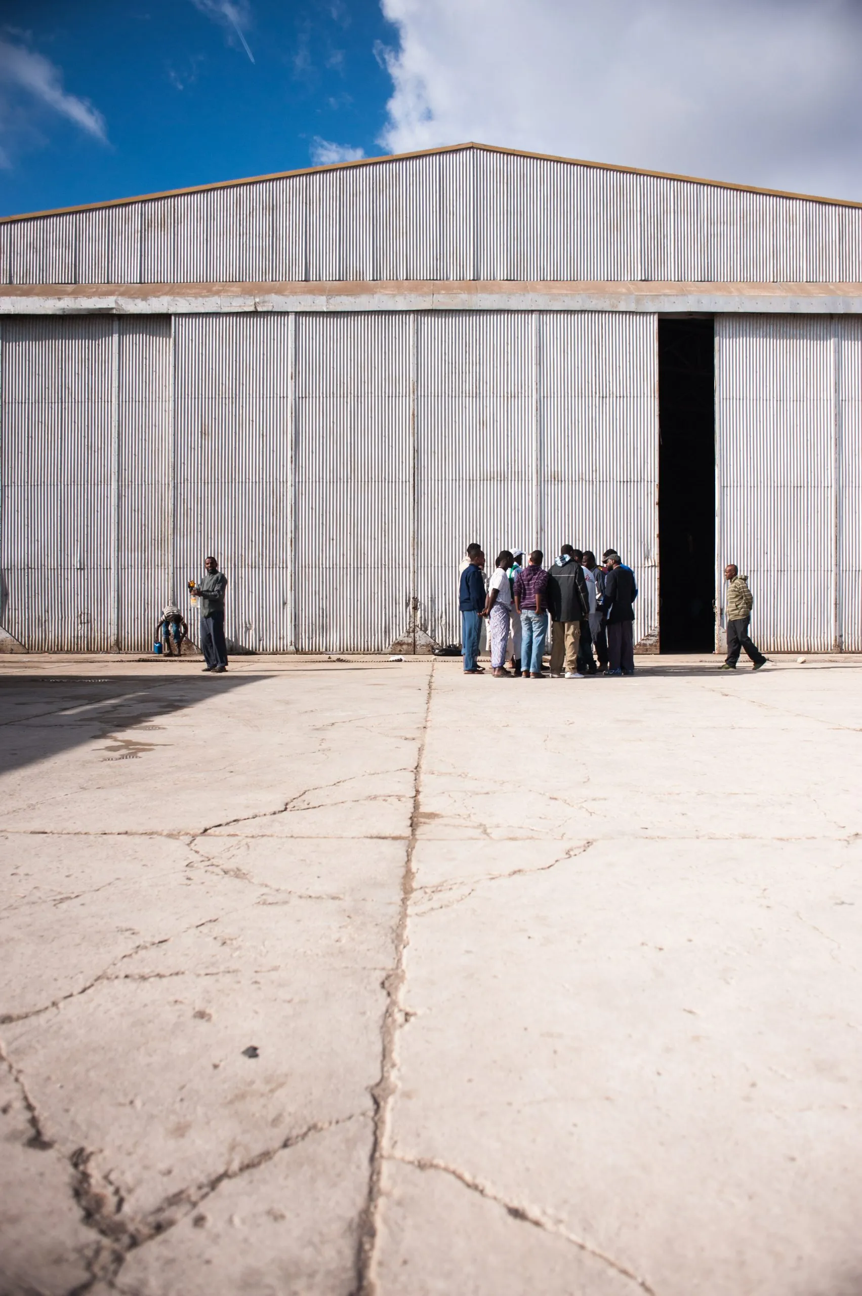 A small group gathers near the entrance of a metal hangar at the Malta asylum compound, under a wide sky.