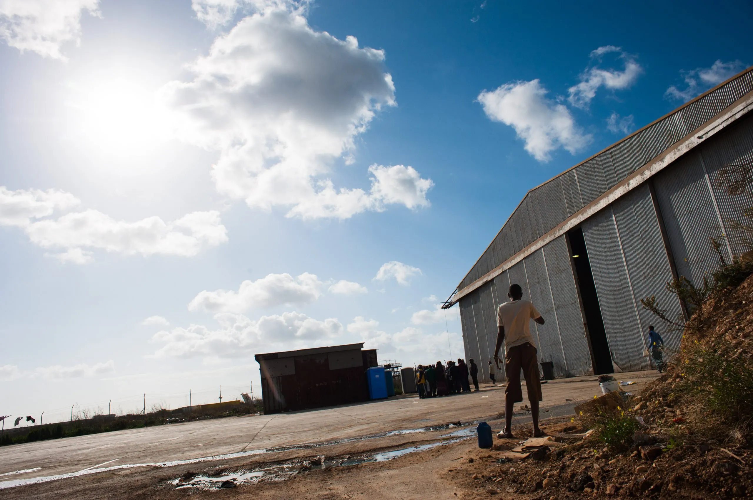 A figure walks past the towering metal facade of the housing hangar at the Malta detention compound.
