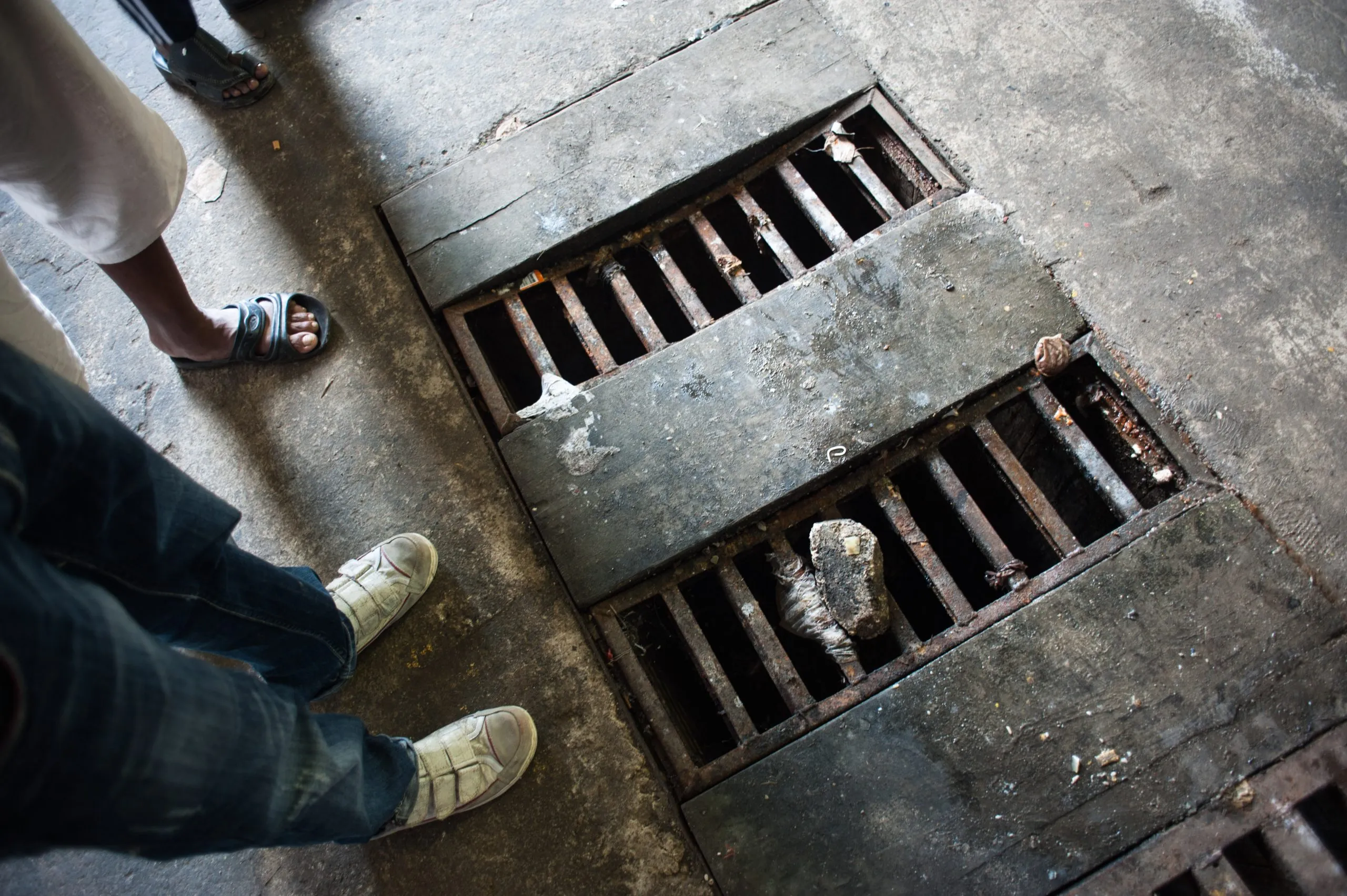 Feet in sandals beside a heavy iron drainage grate on a concrete floor, in the Malta housing hangar.