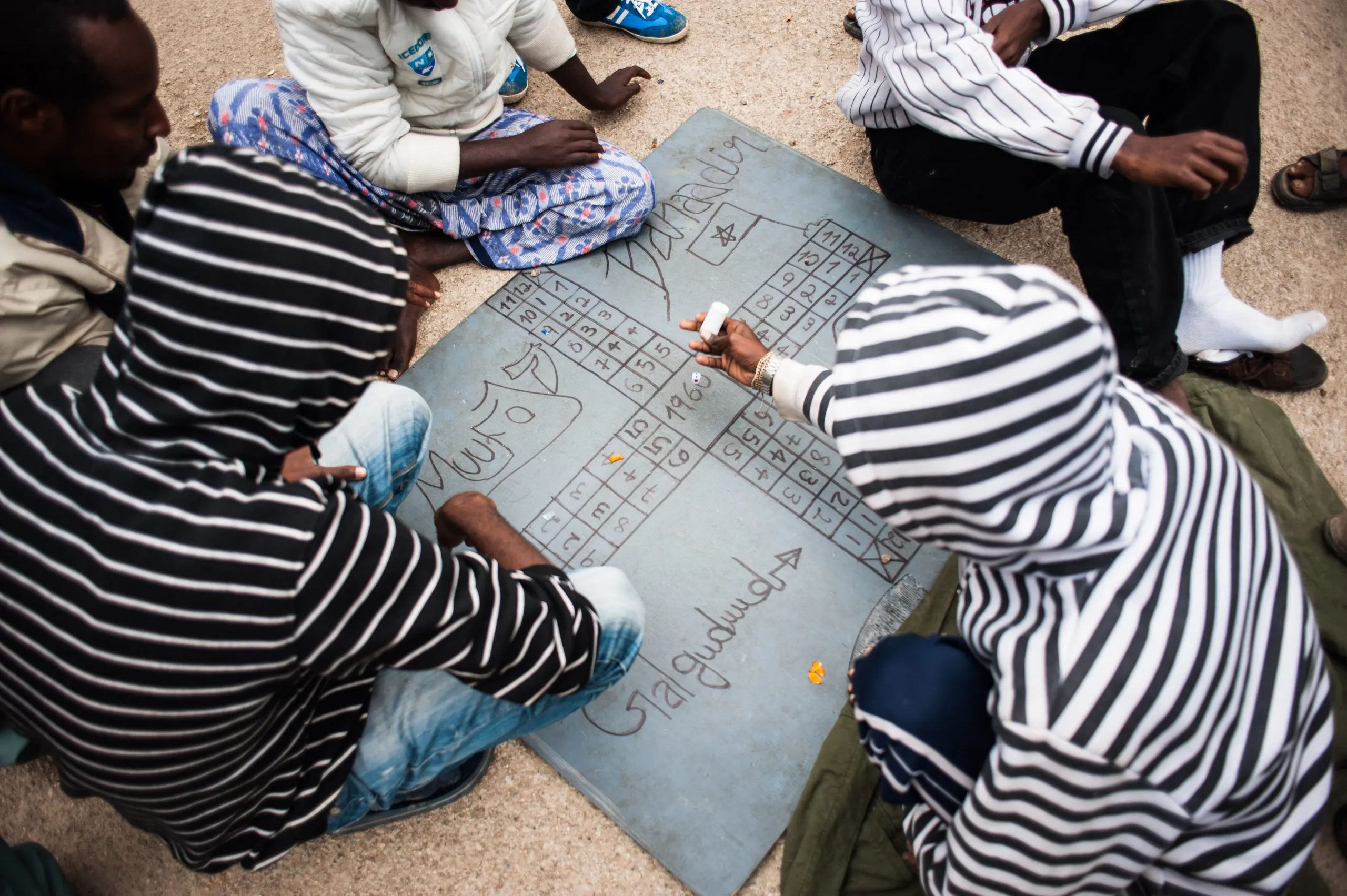 Several people kneel over a sheet of cardboard, drawing and writing together inside the Malta housing compound.