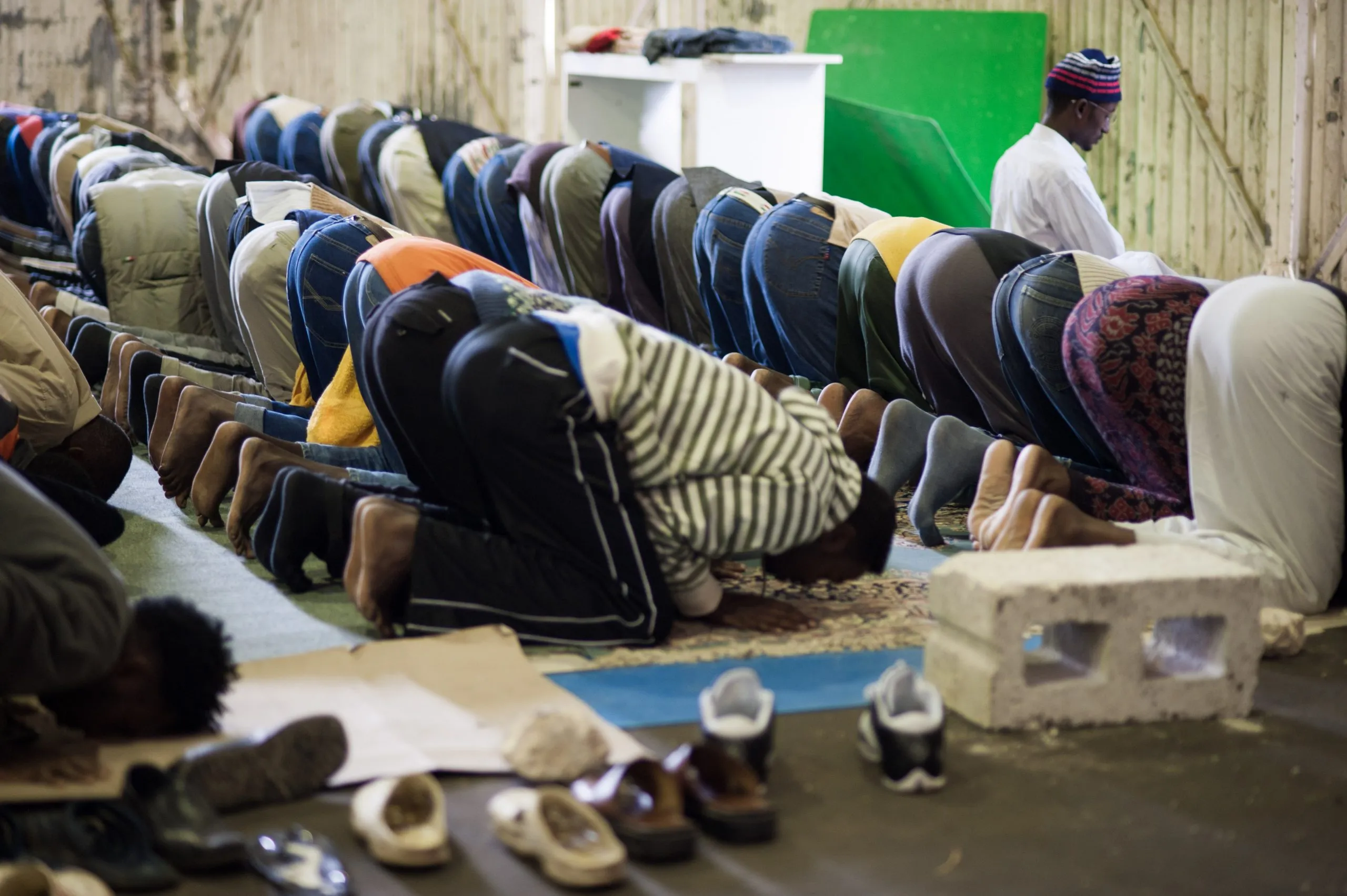 Rows of men in prostration during prayer inside the housing hangar, with shoes lined up at the front.