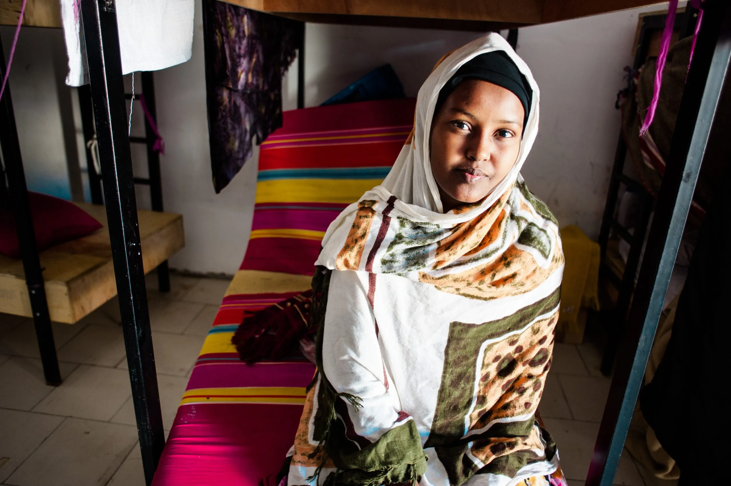 A woman in a headscarf sits on a brightly striped bed in the hangar dormitory on Malta.