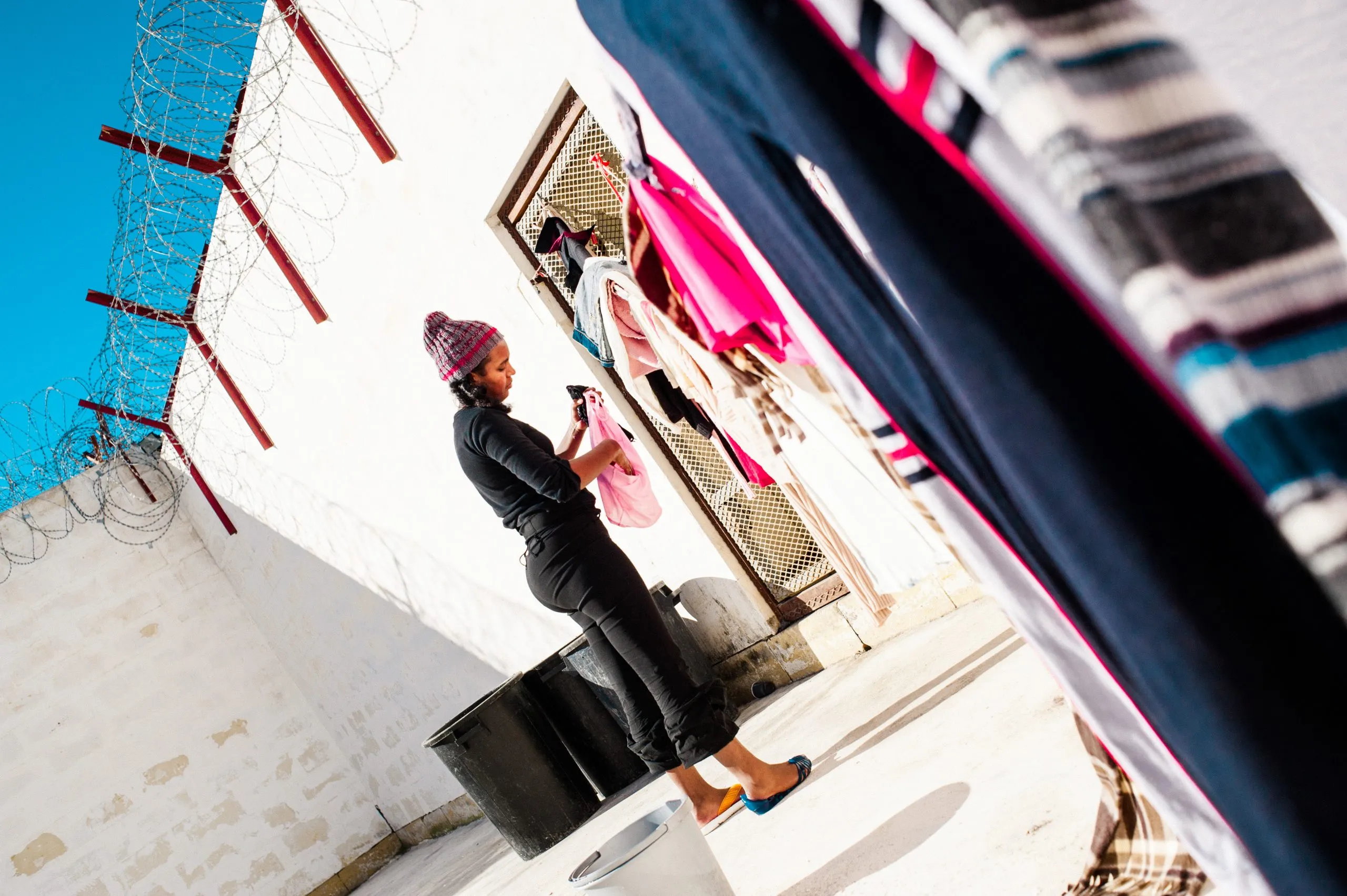 A woman stands on a barrel to hang laundry beside a barbed-wire perimeter wall at the Malta compound.