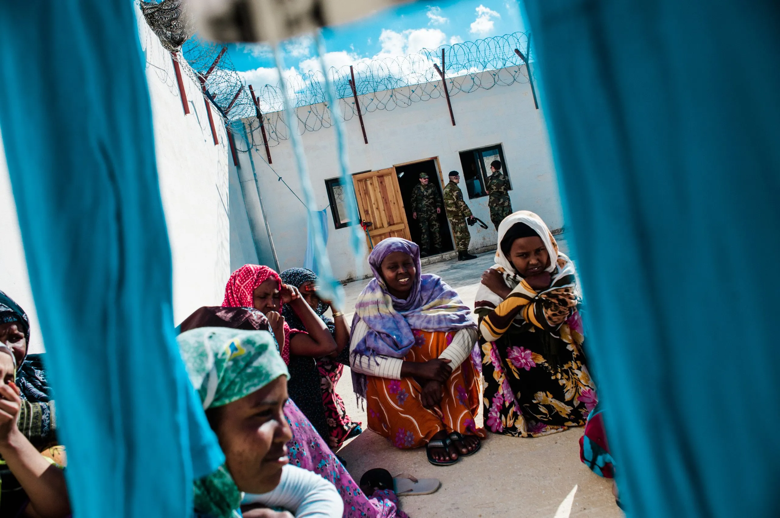 Women in patterned dresses sit in a yard, framed by hanging laundry, with guards beyond the barbed-wire wall.