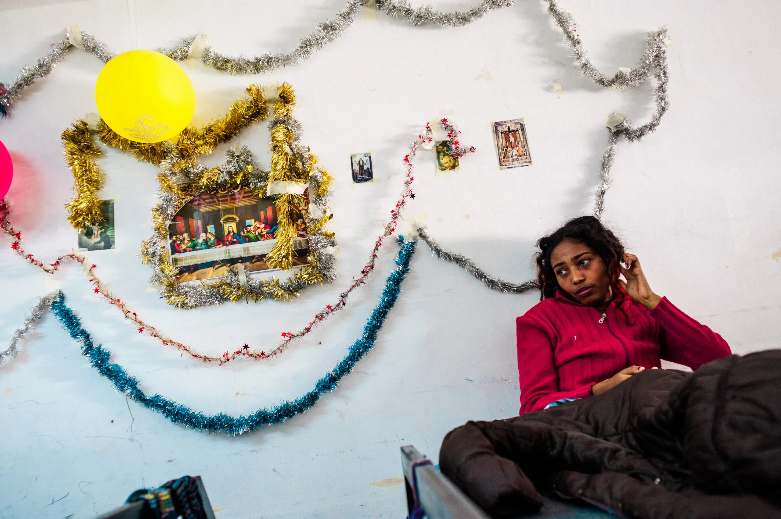 A woman in a red top rests in a room hung with Christmas tinsel and small printed icons, on Malta.