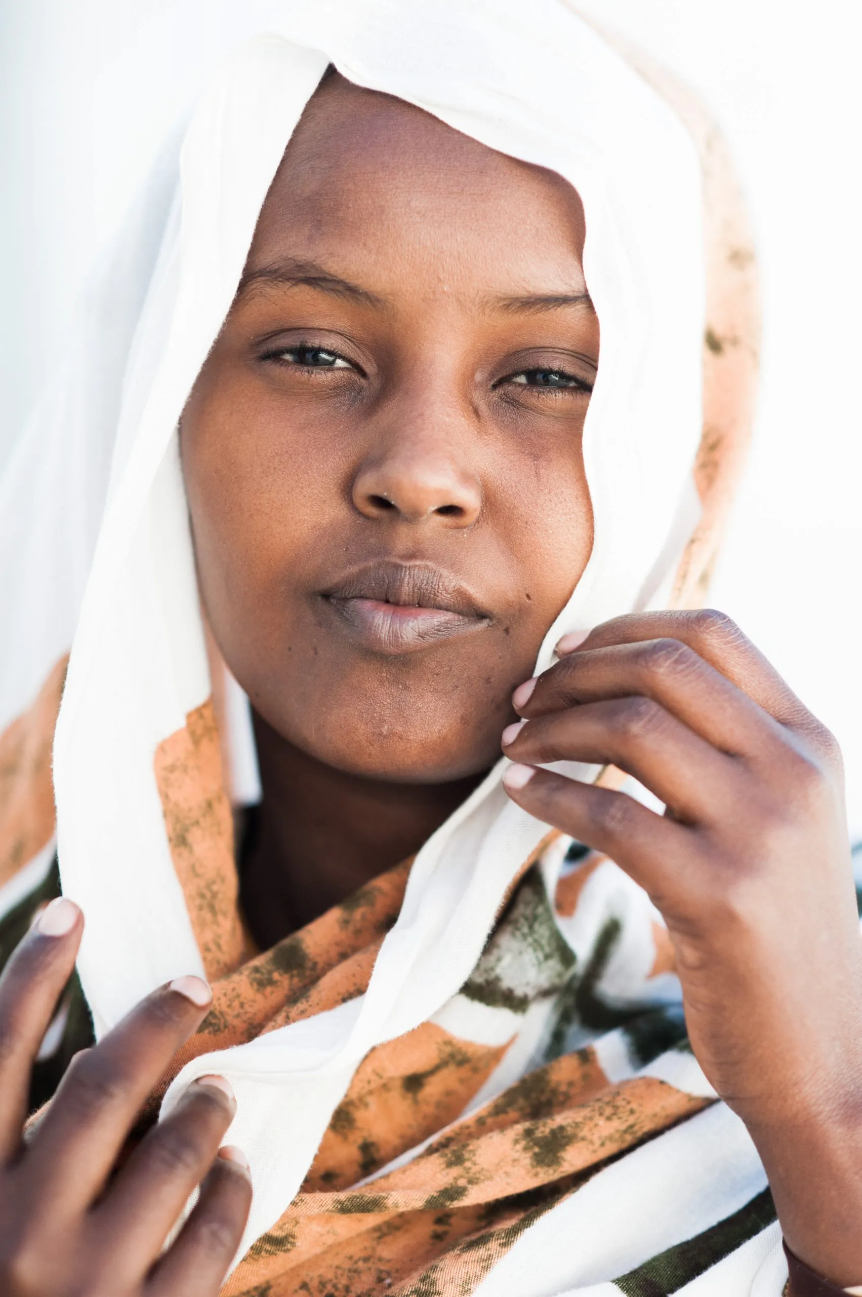 A close portrait of a young woman in a patterned white-and-orange headscarf, against bright daylight.