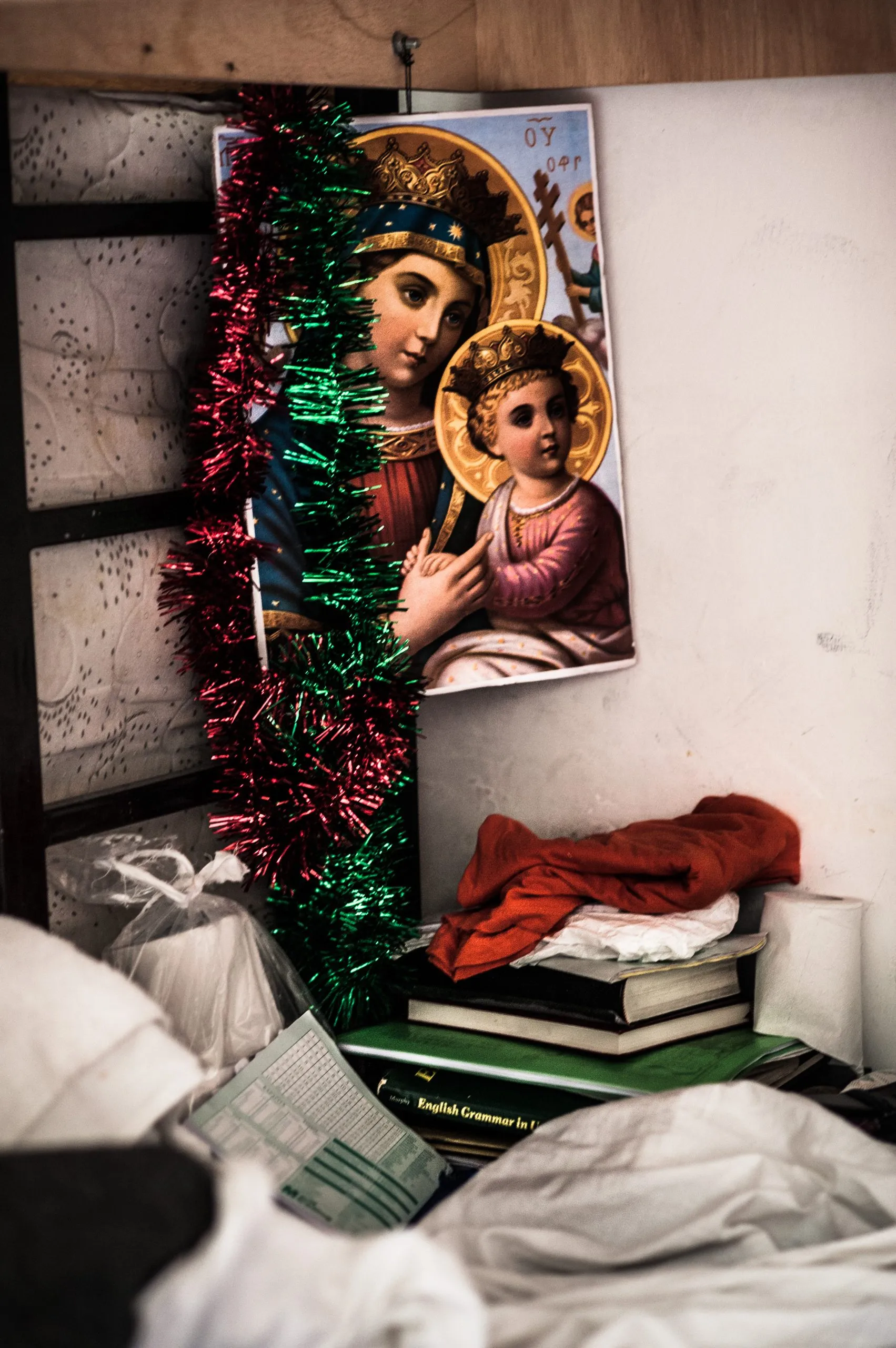 A printed Madonna and Child icon framed by Christmas tinsel above a stack of books inside the housing compound.