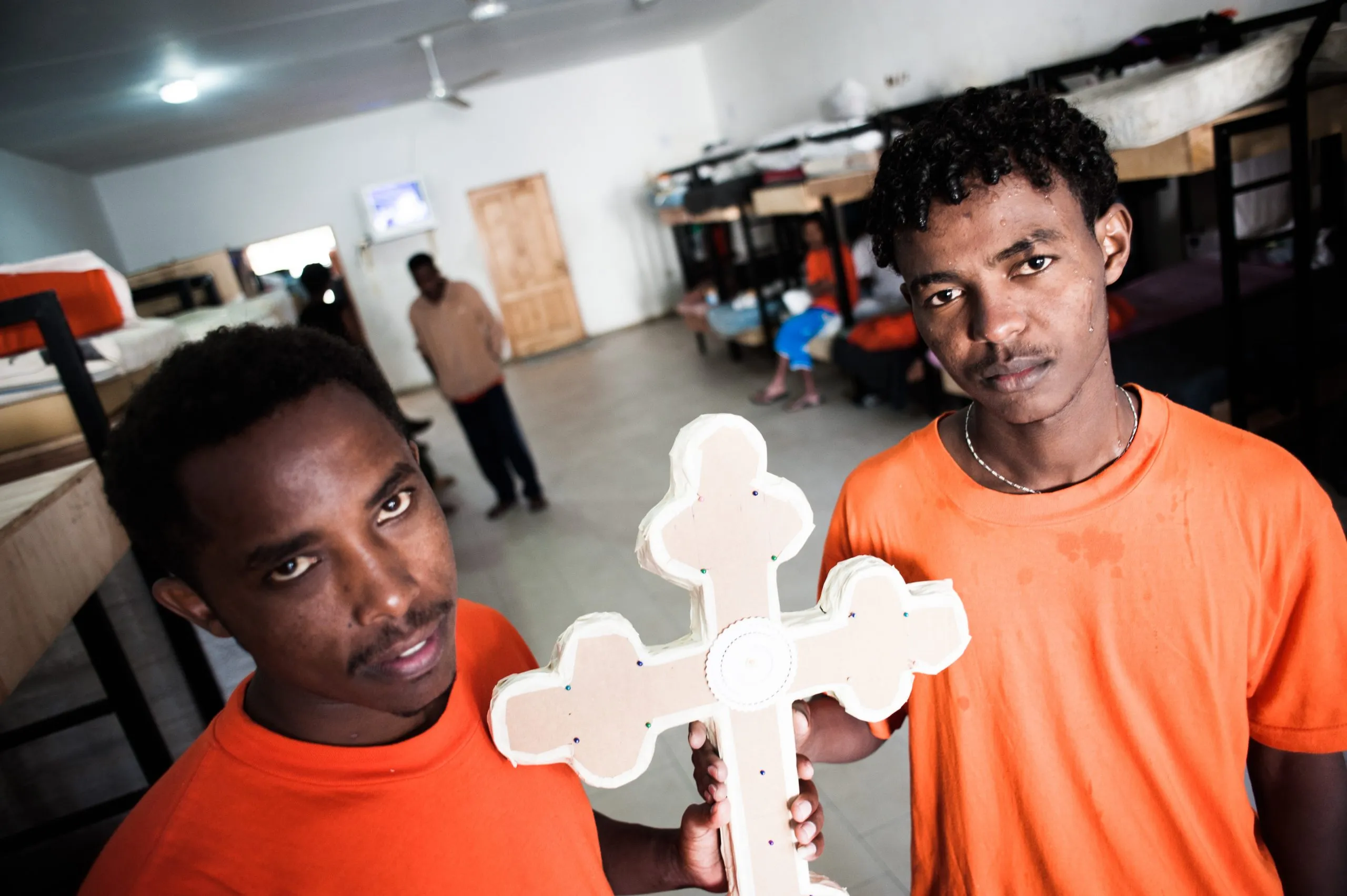 Two men in orange shirts hold a white wooden cross inside the dormitory hangar on Malta.