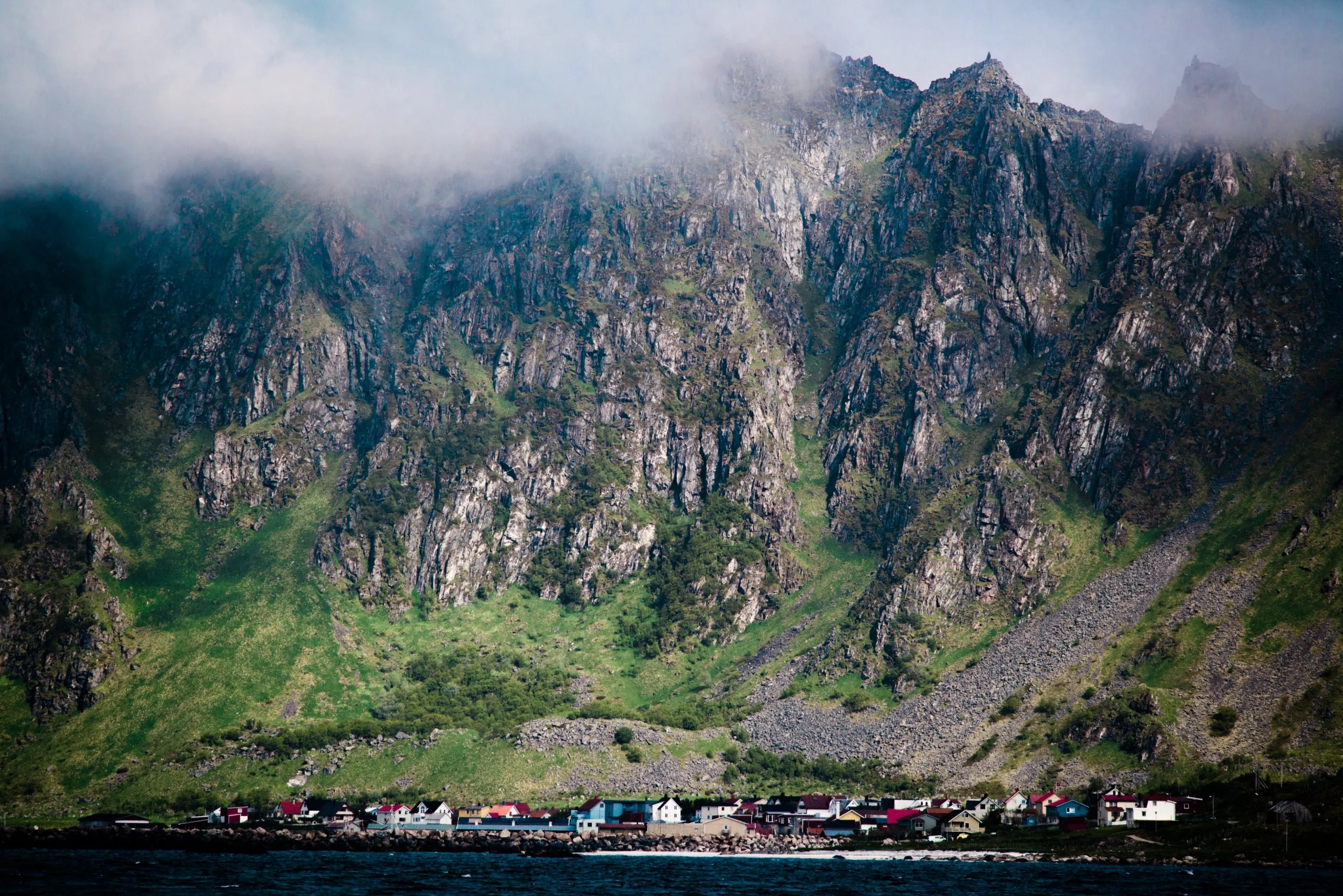 A small Norwegian fishing village hugs the shore beneath the cloud-wrapped granite walls of a Lofoten / Vesterålen mountain.