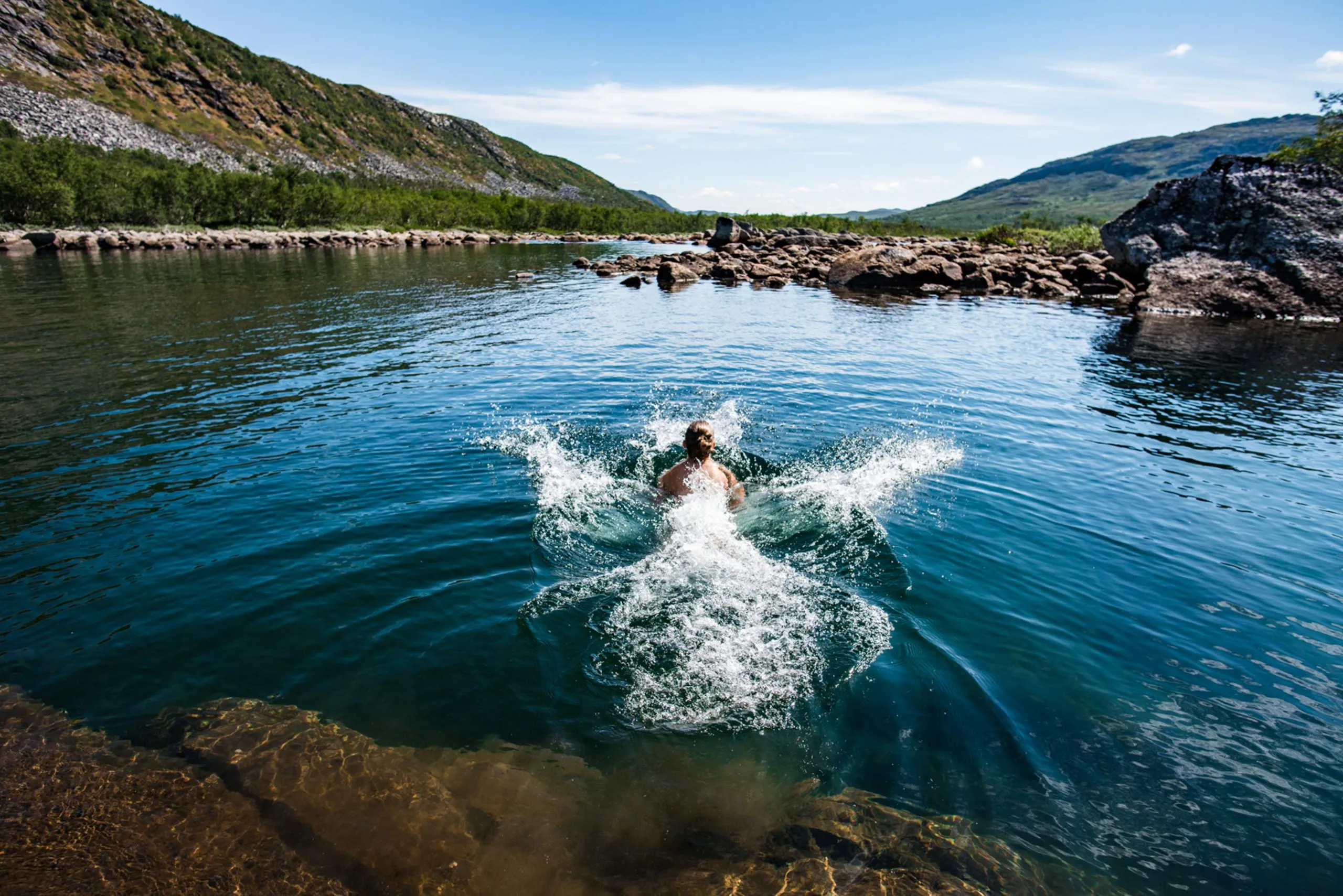 A swimmer dives into the clear water of a mountain river on Norway's Hardangervidda plateau, splash radiating outward in midday sun.