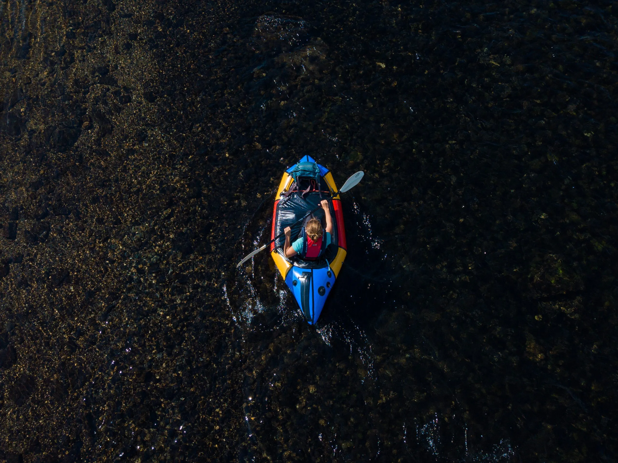 Drone view of a person paddling an inflatable kayak through dark seaweed-covered shallows on the Norwegian coast.