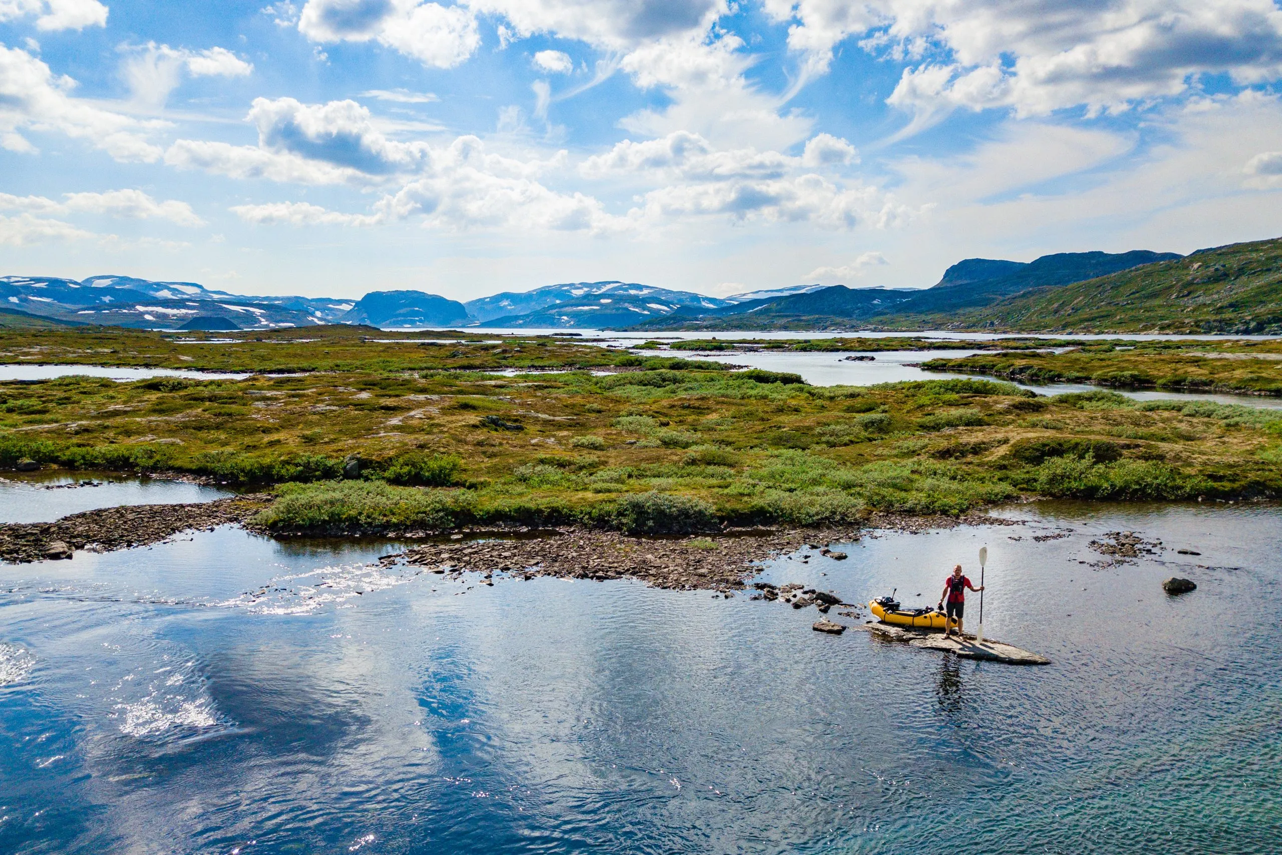 Aerial view of a person on an inflatable packraft drifting through a maze of mossy islets and clear water on Norway's Hardangervidda plateau.