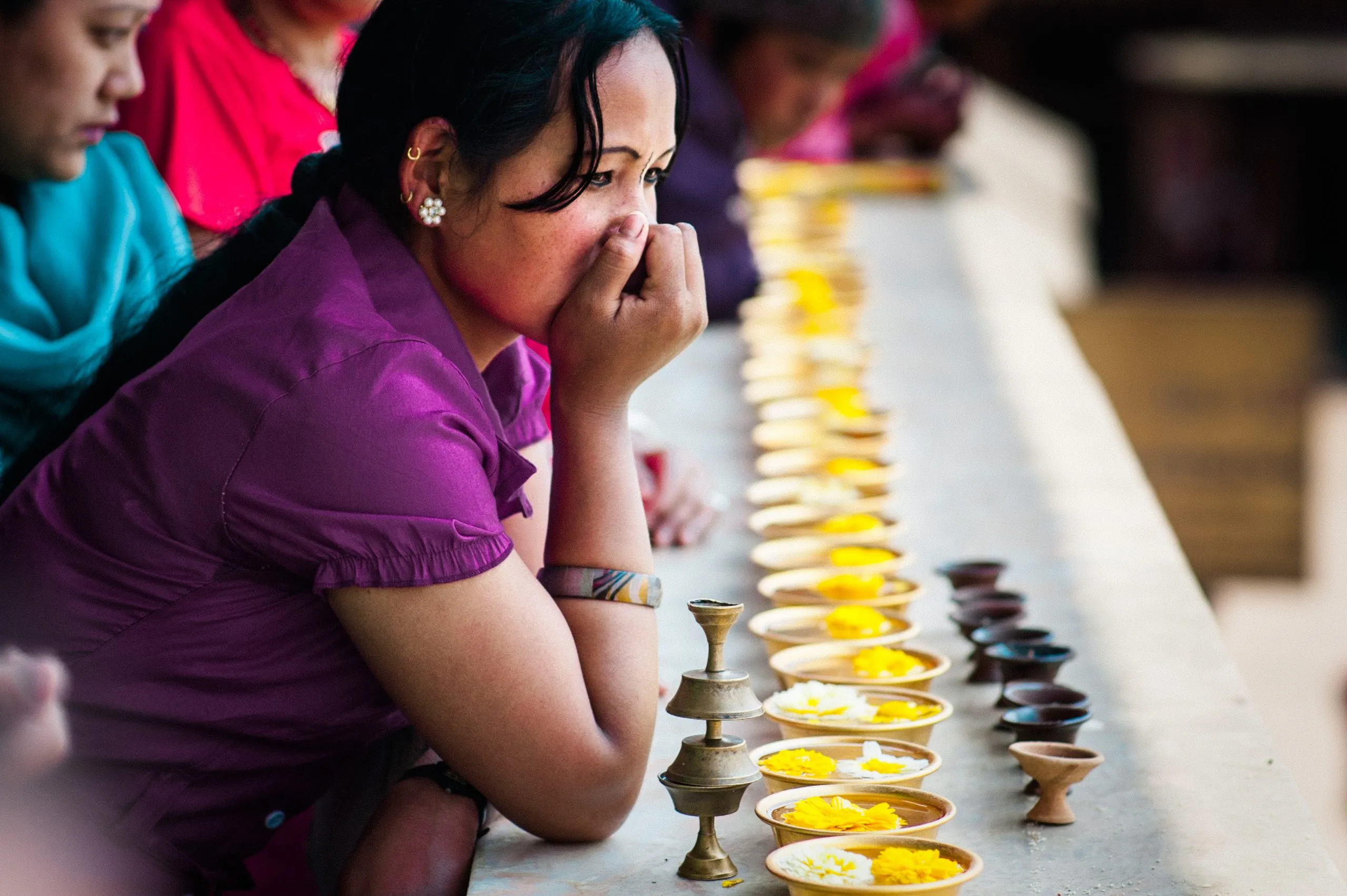 A Nepali woman in a purple top leans on a long stone counter holding rows of small clay butter-lamp cups topped with marigolds at Pashupatinath.