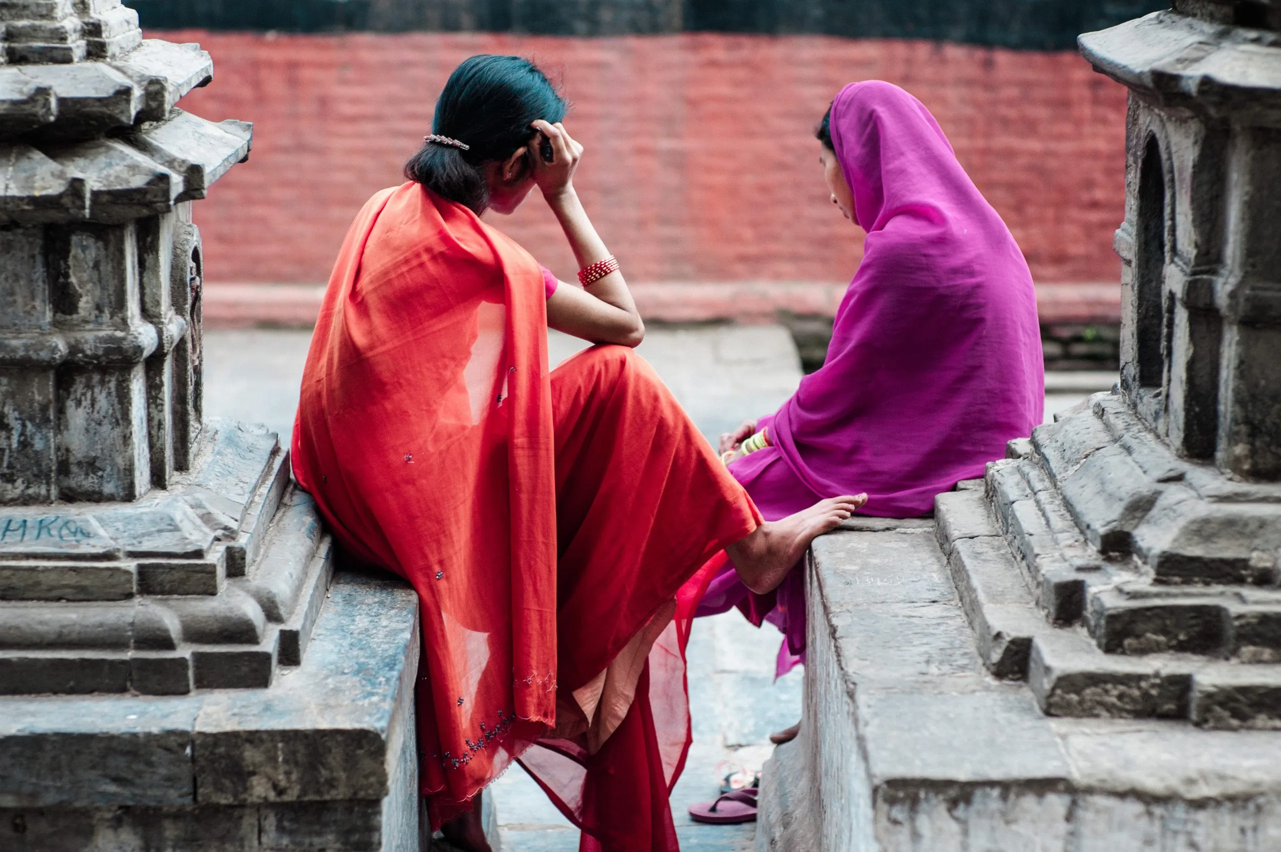 Two Hindu women in a red and a magenta sari sit on the stone steps of Pashupatinath temple in Kathmandu, deep in conversation.