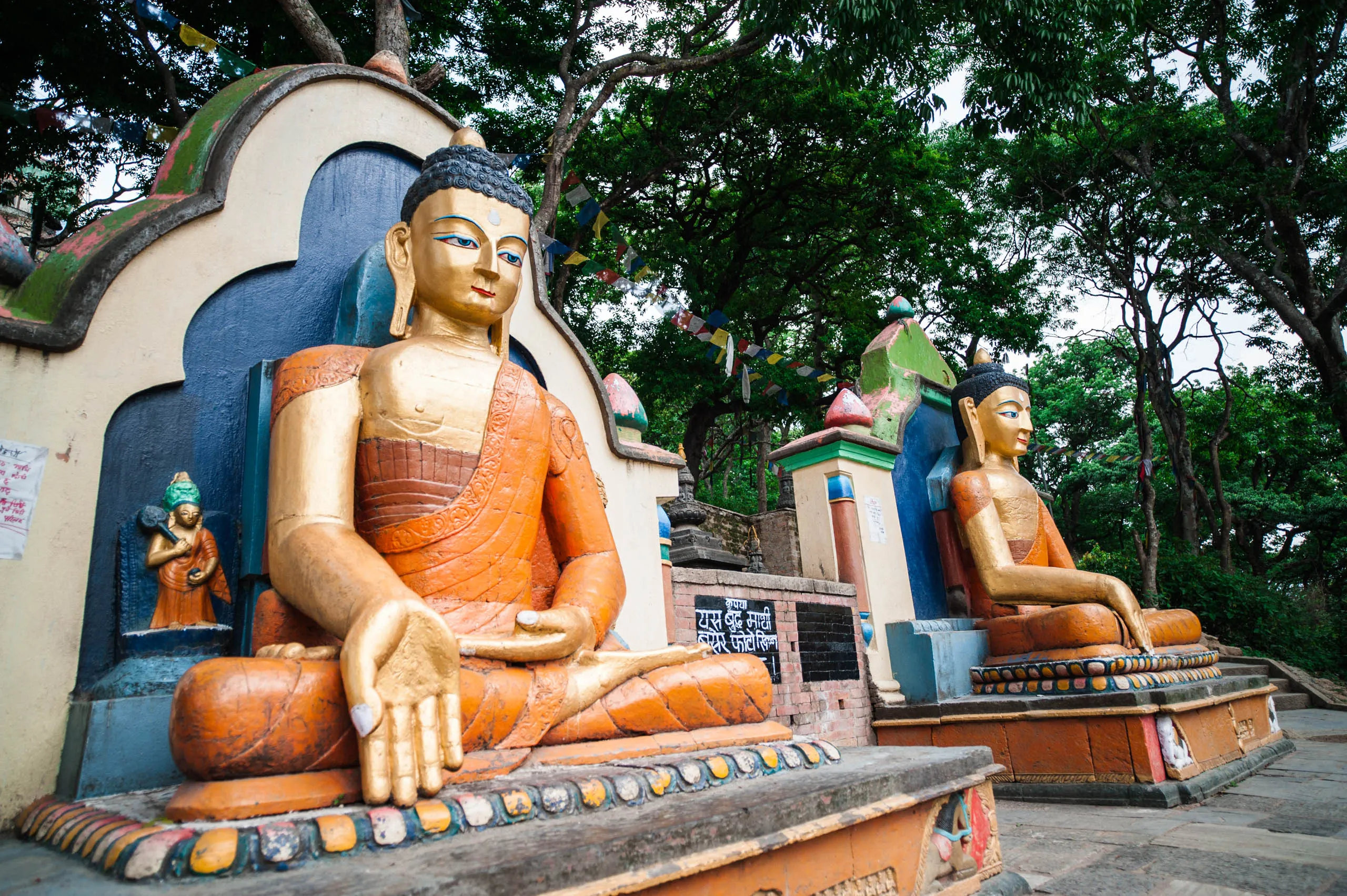 Two large gilded Buddha statues seated at the base of Swayambhunath temple in Kathmandu, beneath prayer flags.