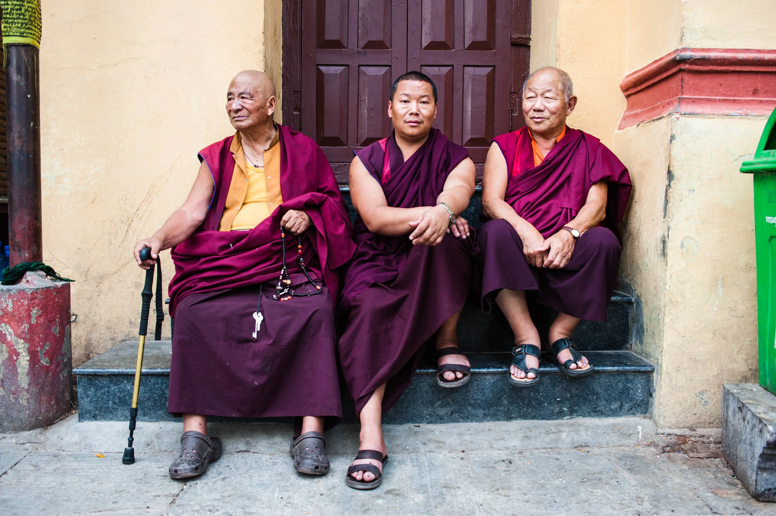 Three older monks in maroon robes sit on a bench at the doorway of the monastery at Swayambhunath, Kathmandu.