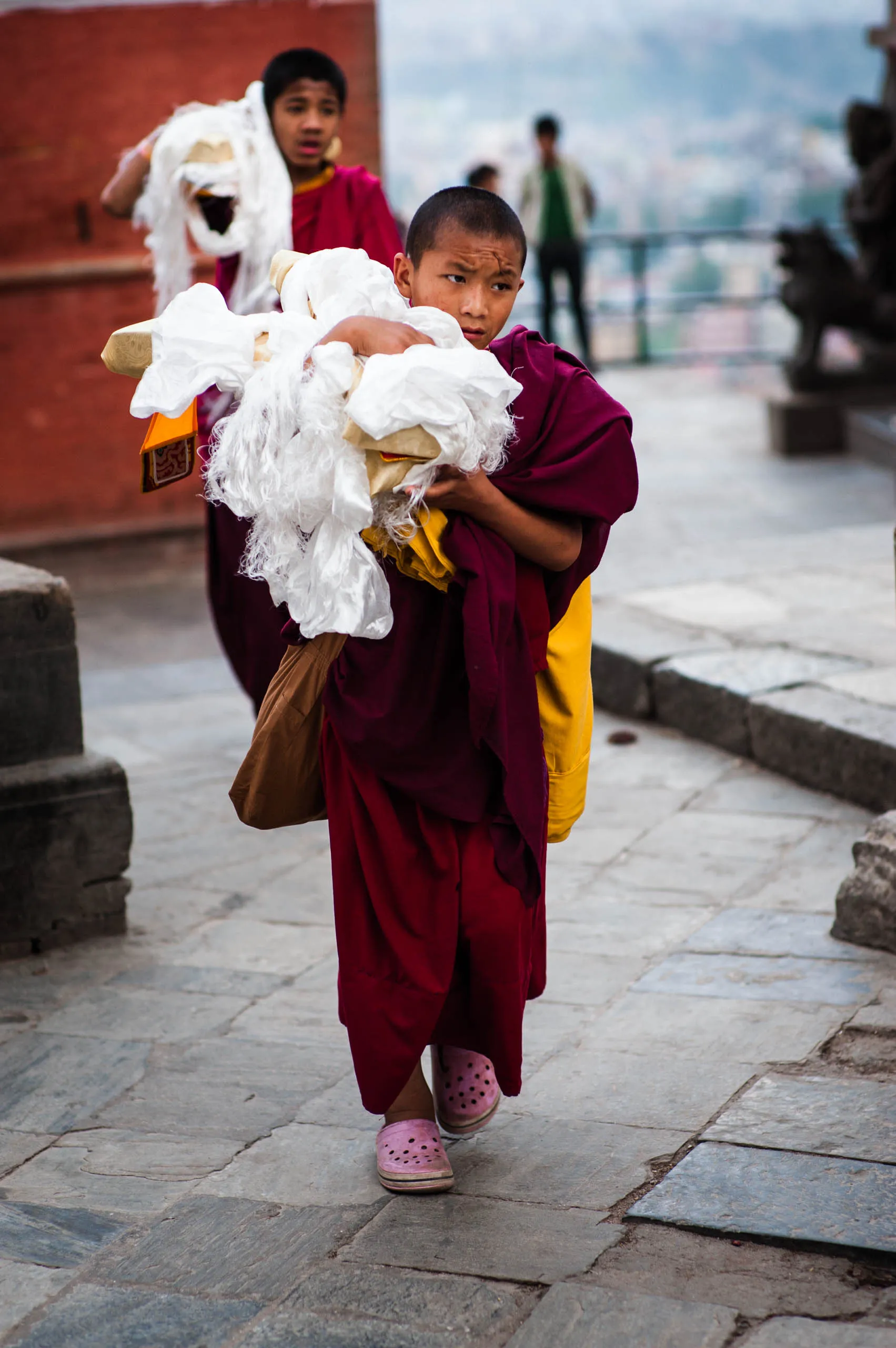 A boy monk walks across the stone courtyard at Swayambhunath, his sash slung over one shoulder.