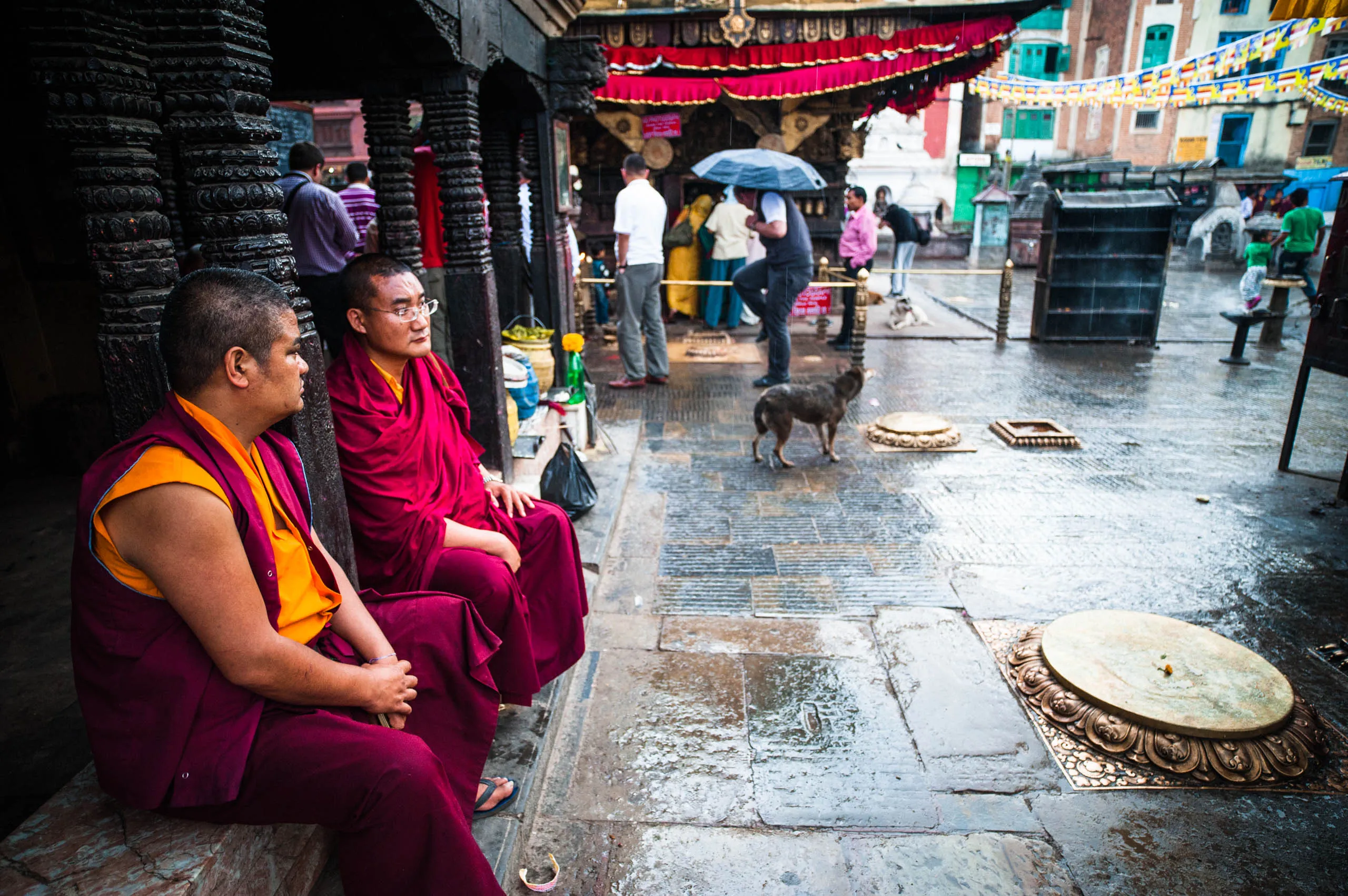 Two monks rest under the painted eaves of the temple courtyard at Swayambhunath, with a stray dog nearby.