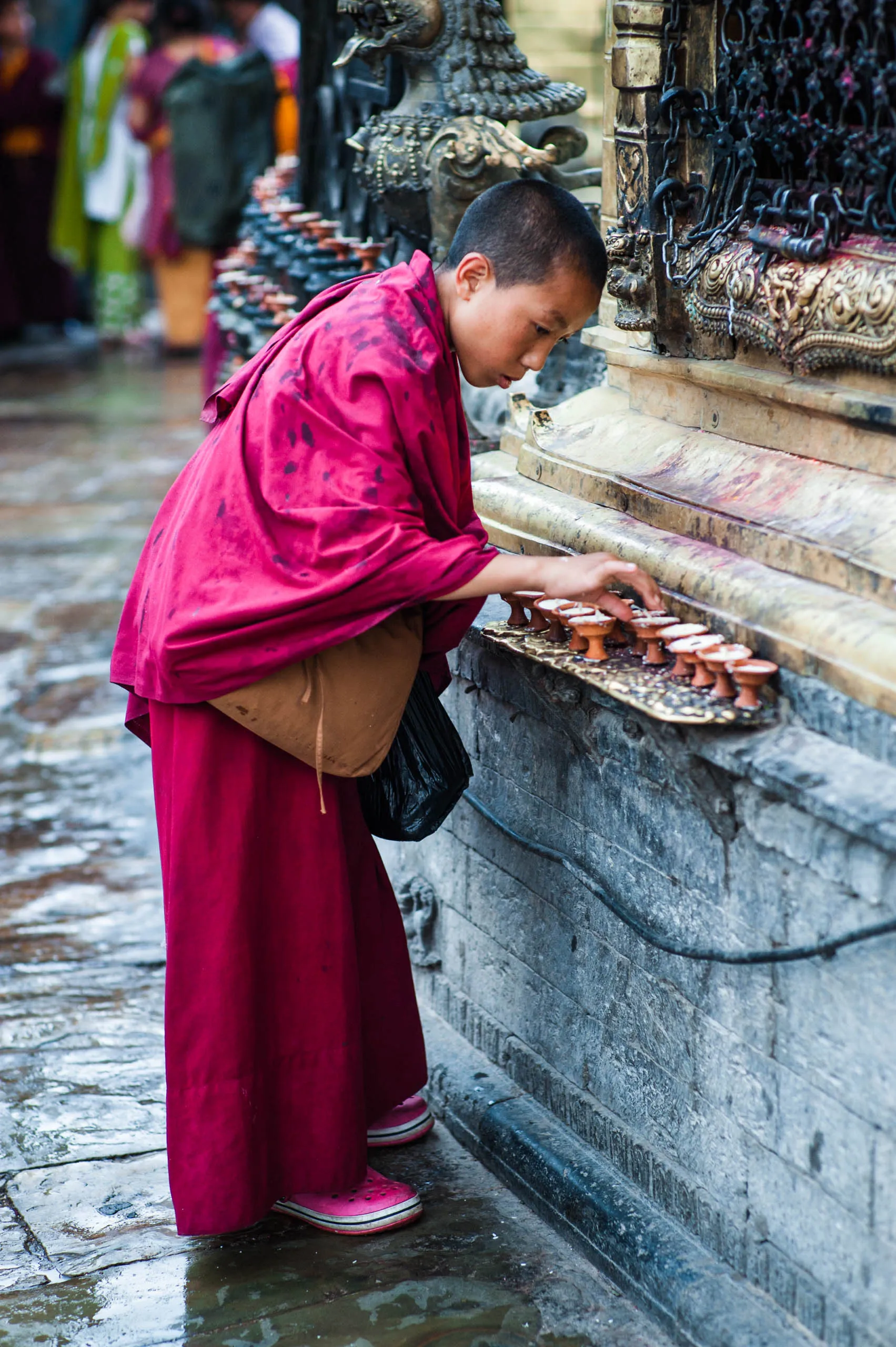 A boy monk leans over a row of small clay butter lamps placed against a stone wall at Swayambhunath.