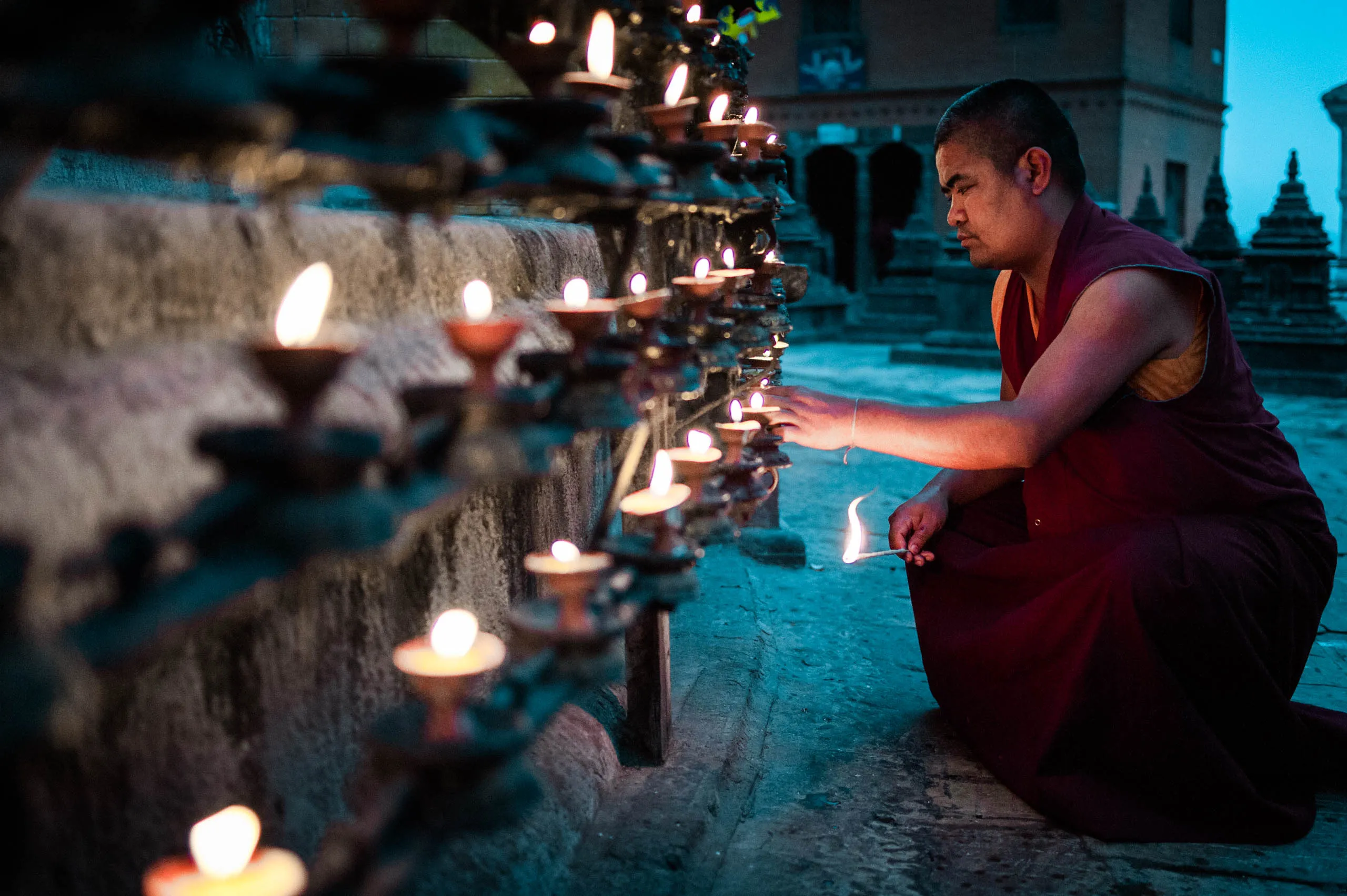 A single monk crouches by a row of butter lamps, lighting them one by one at Swayambhunath.