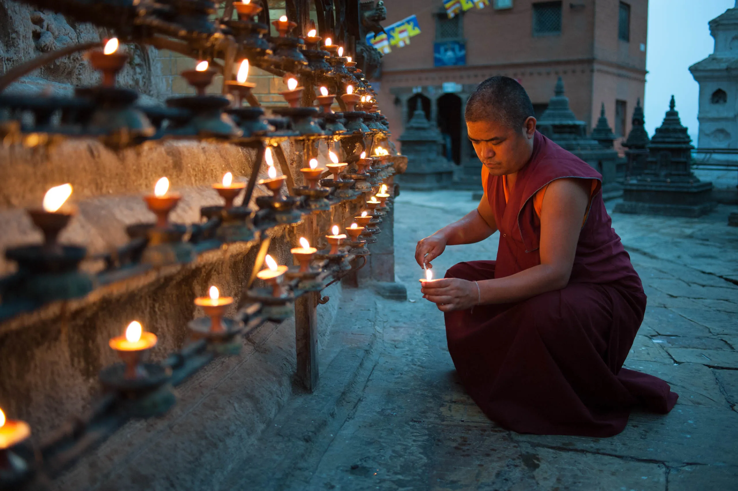 A monk lights a butter lamp on the iron railing surrounding a small stupa at Swayambhunath, at dusk.