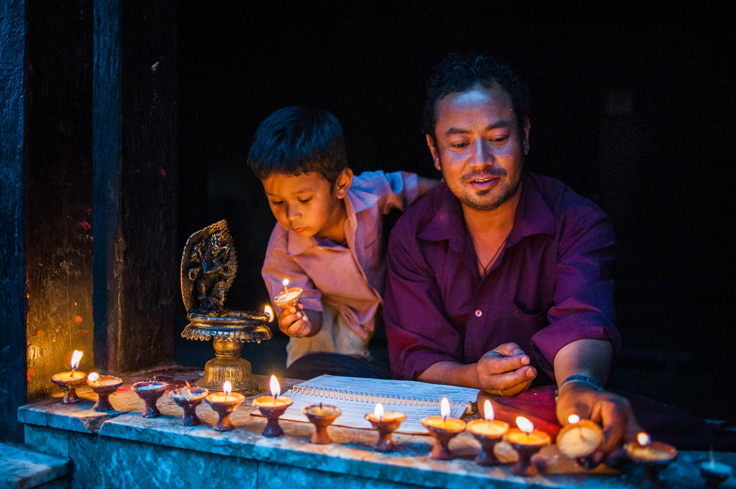 A man and a small child arrange and light butter lamps from an instruction book at Swayambhunath temple.