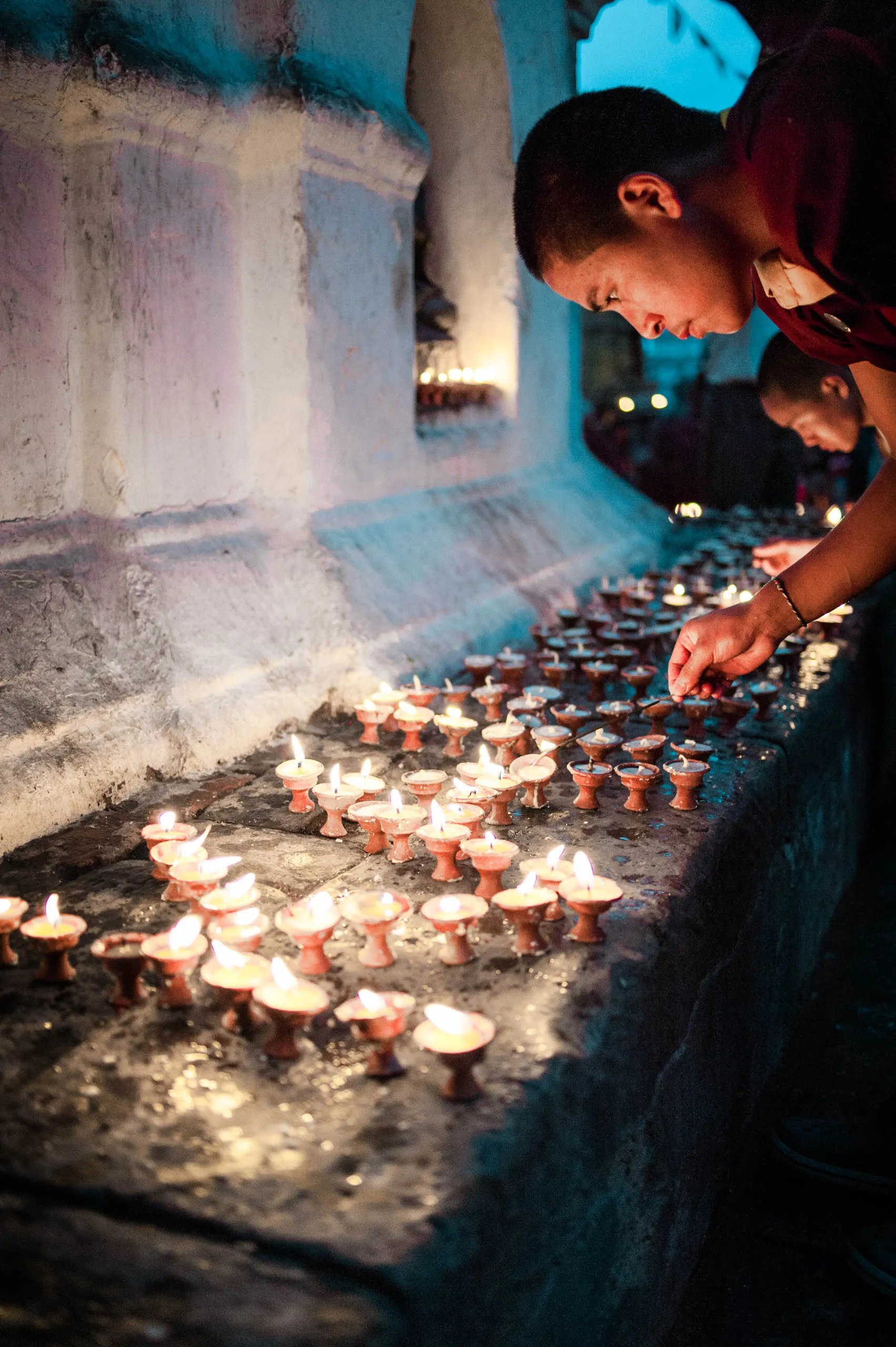 A monk leans across a stone ledge to light a long row of butter lamps along the side of a stupa.